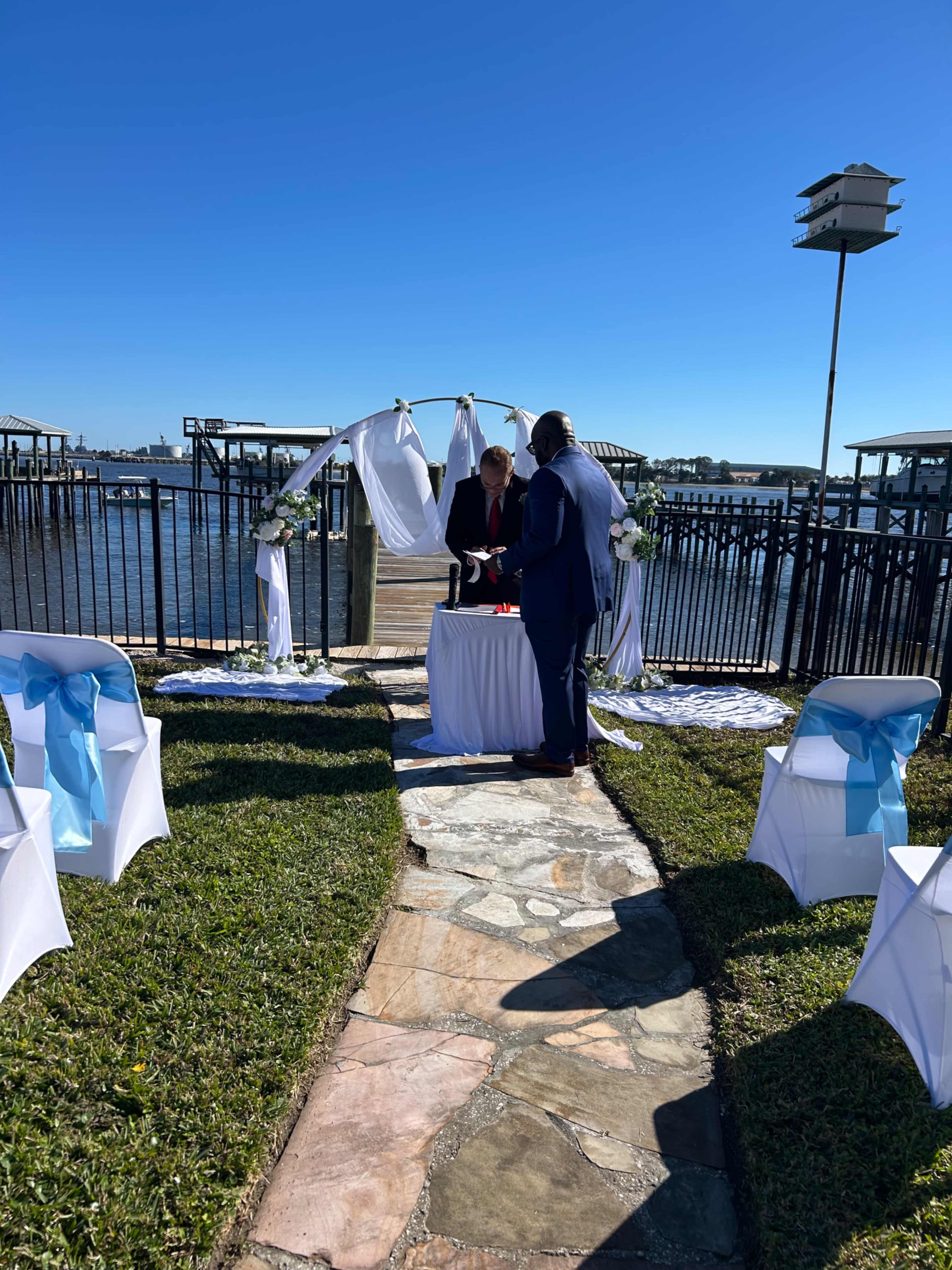 A couple stands at an altar decorated with white drapery by a body of water, surrounded by white chairs with blue sashes.