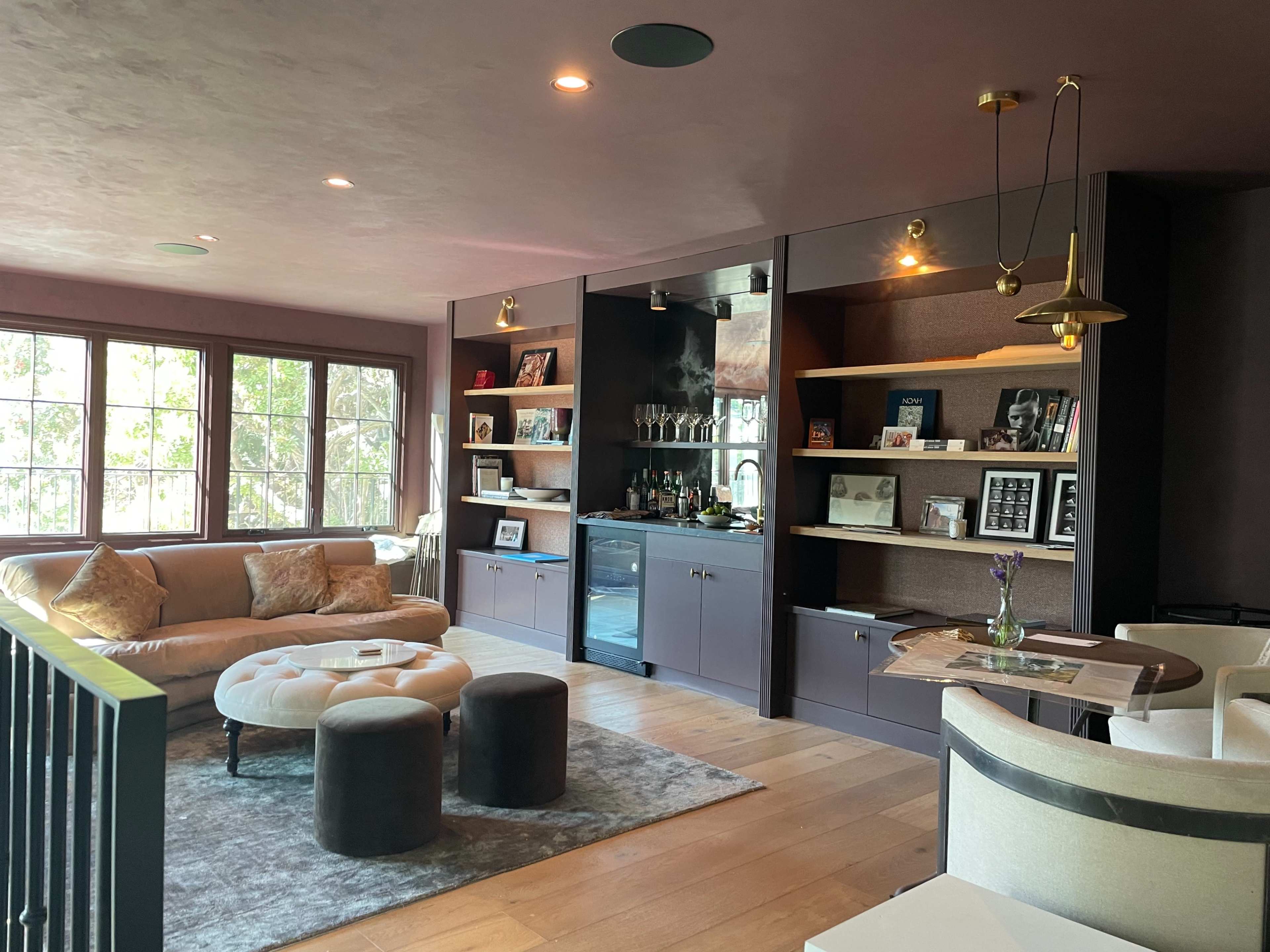 The image shows a modern living room with a sofa, a round coffee table, shelves containing books and decor, and a bar area with a beverage cooler, all featuring large windows that let in natural light.