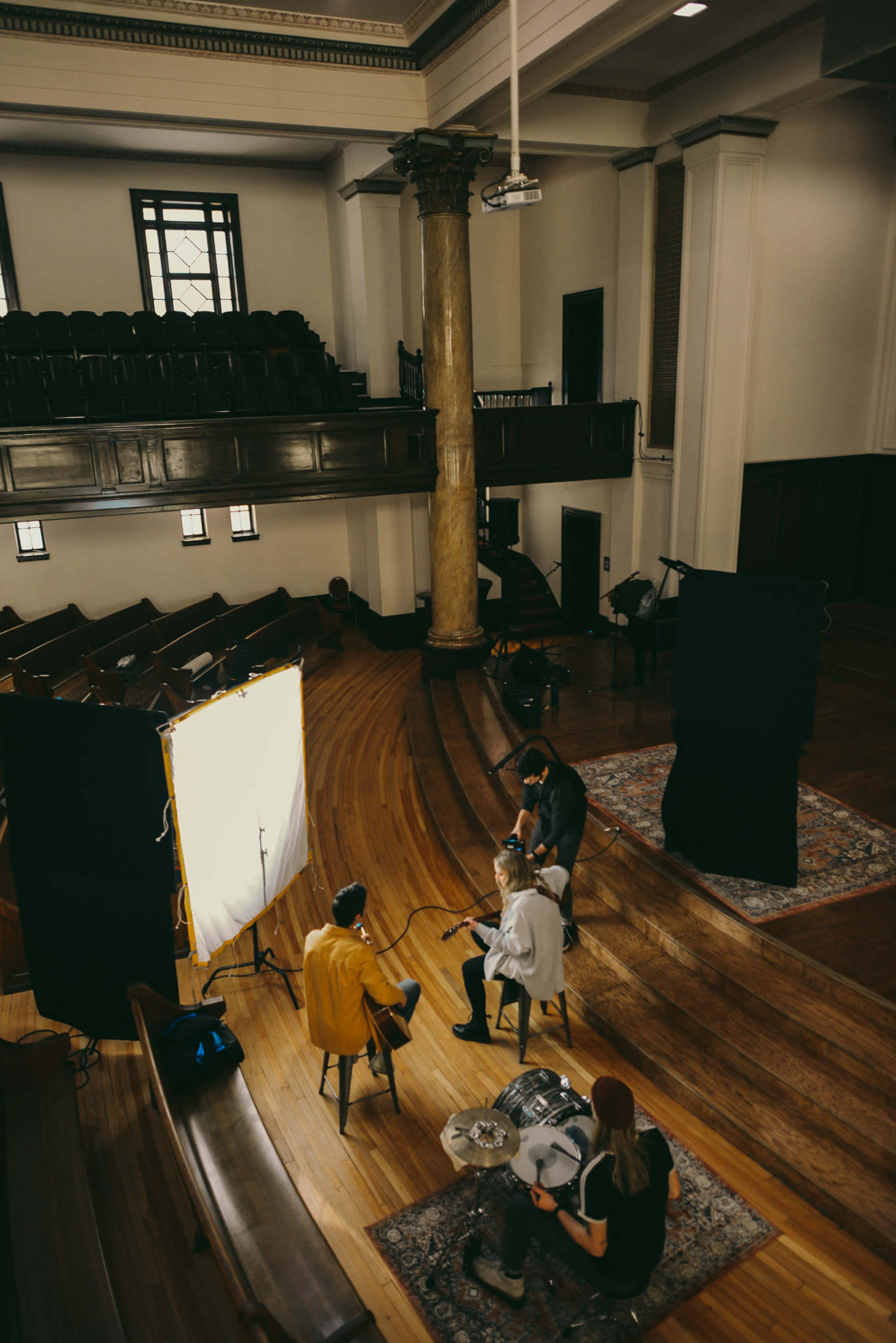 A small film crew sets up in a hall with wooden flooring and a raised seating area, featuring three people seated on stools facing bright lighting and cameras.