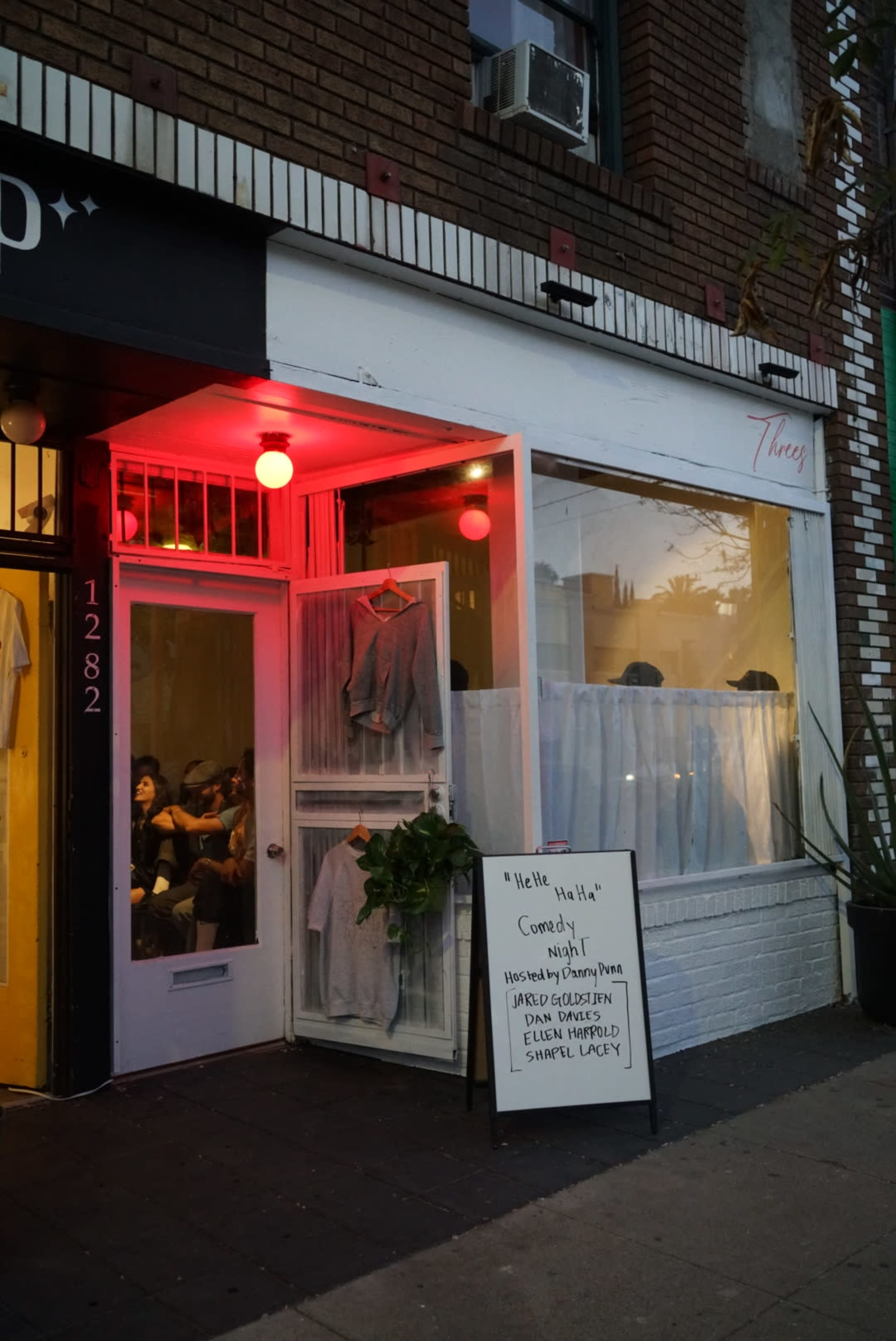 A restaurant entrance with a white facade, a menu sign, potted plants, and people visible inside.