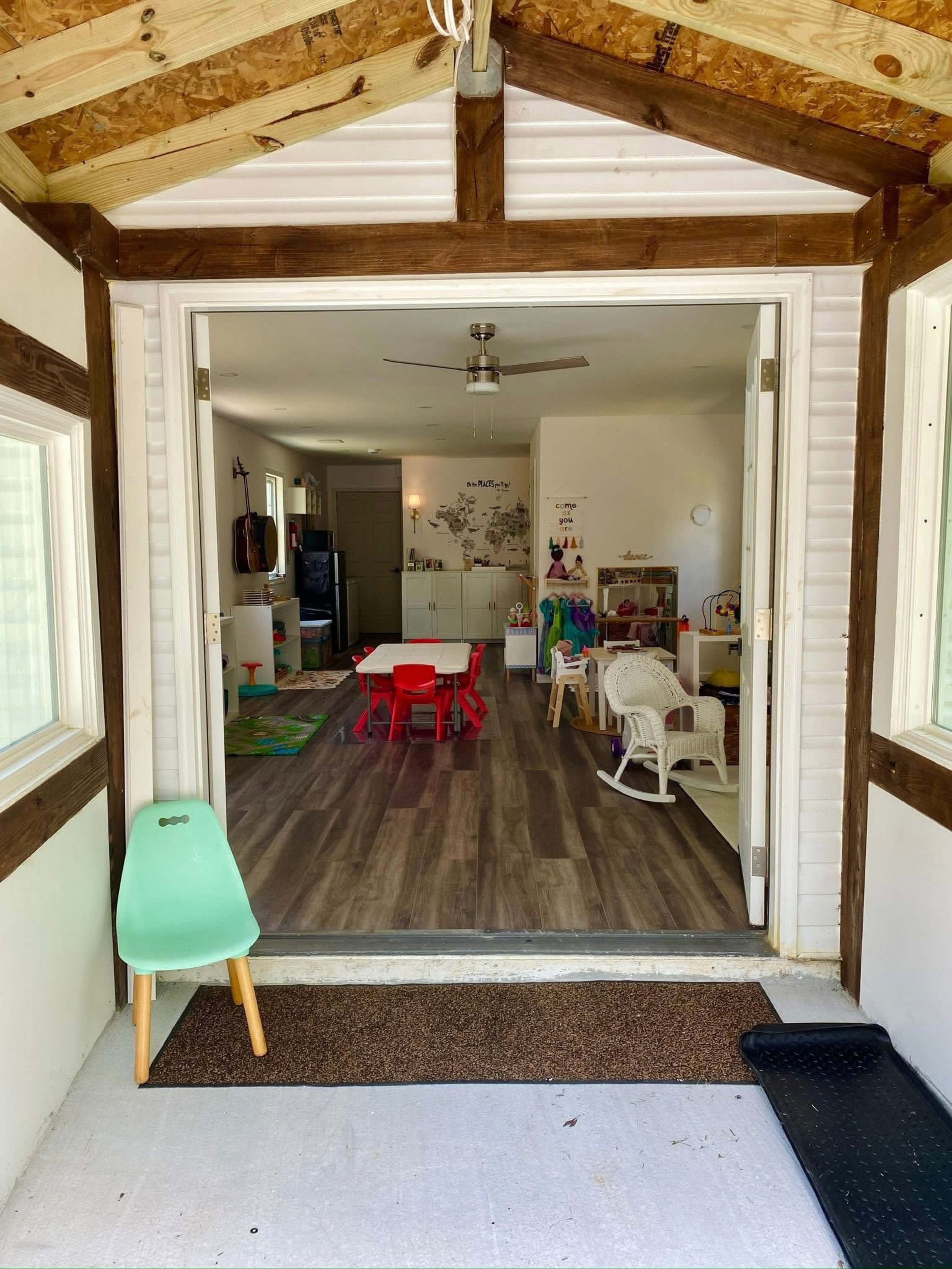 The image shows a view from an entrance into a brightly lit playroom with a table and chairs, toys, and a kitchen area visible in the background.