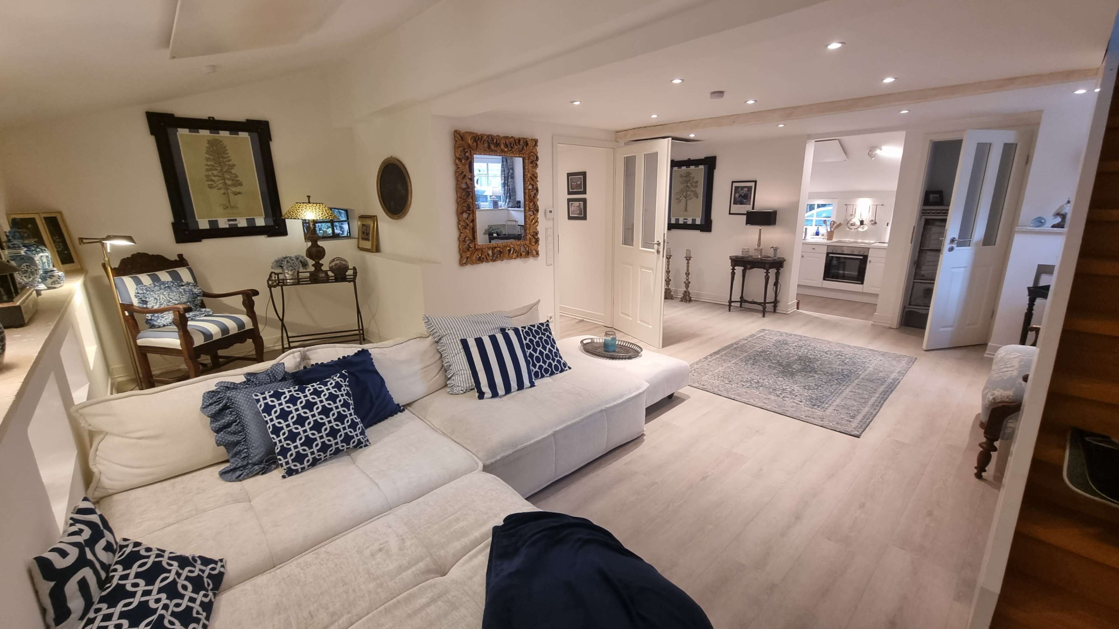The image shows a modern living room with a white sectional sofa, decorative pillows, and an open layout leading to a bright kitchen area.