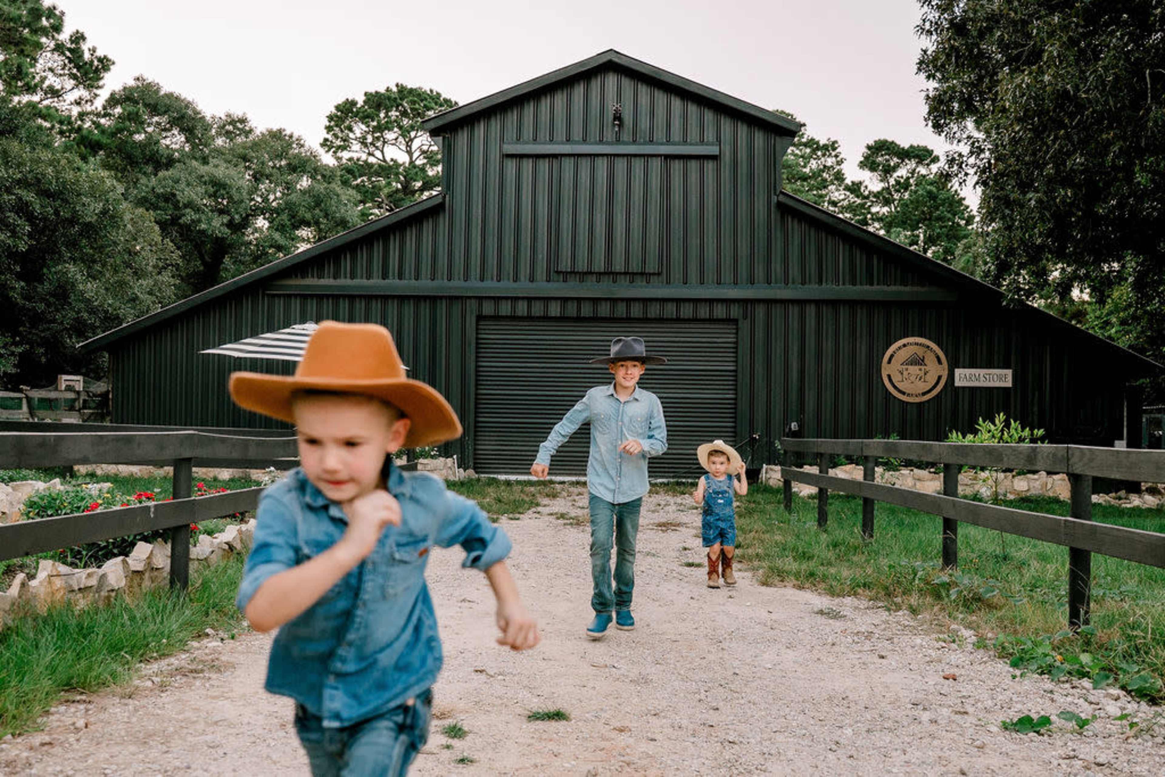 Three children wearing hats run along a dirt path towards a large green barn in a rural setting.