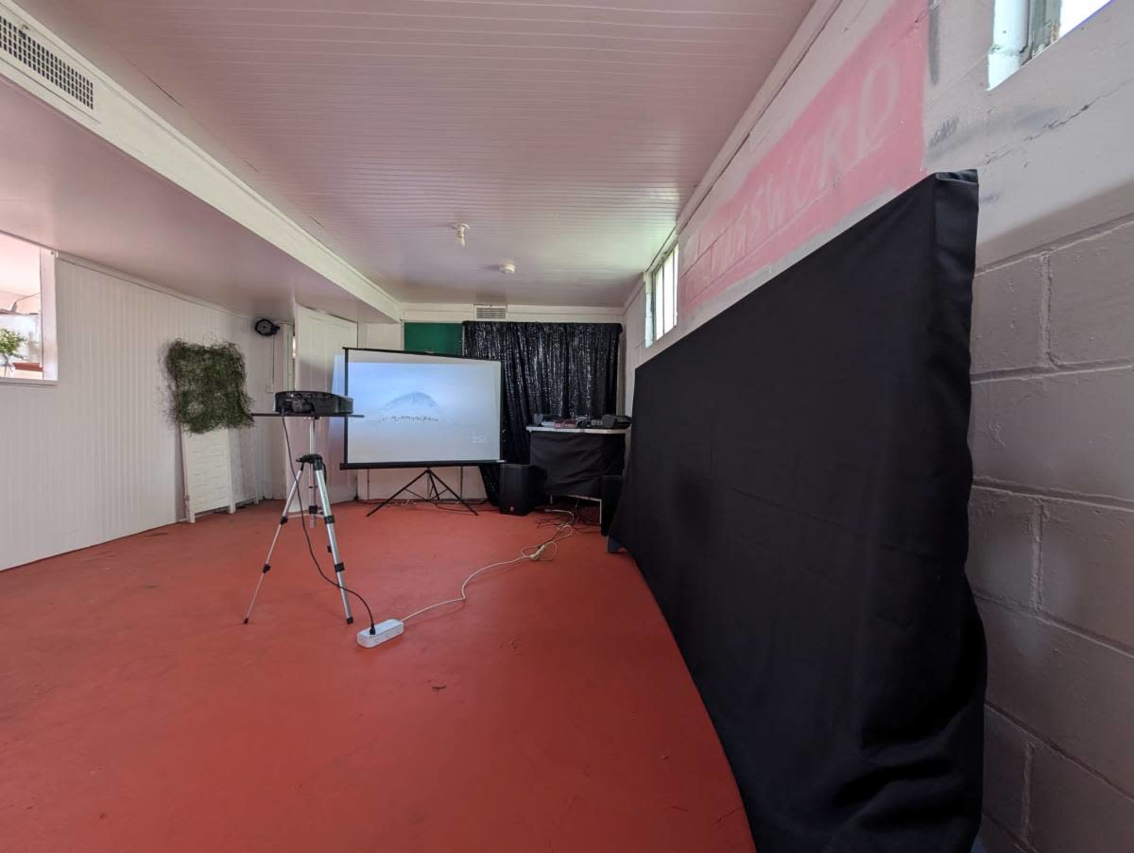 The image shows a small indoor space with a red floor, featuring a projector screen, a tripod, and a black backdrop.