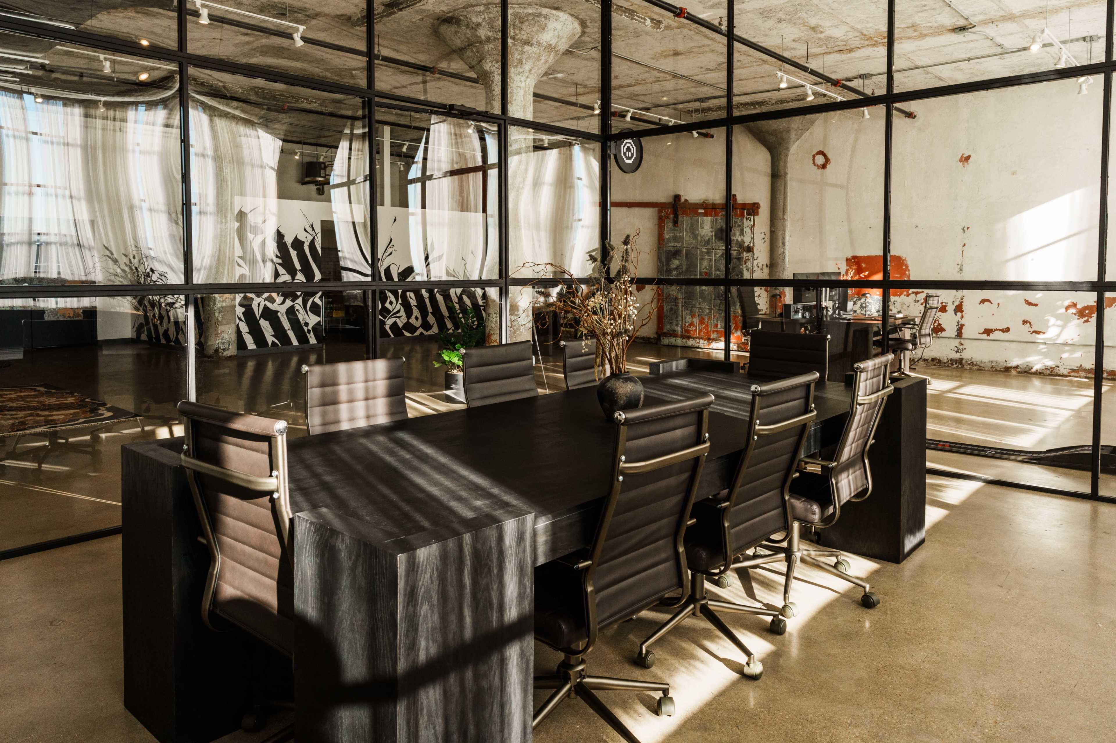 A large conference room with a dark, rectangular table and several black chairs is surrounded by glass walls, showcasing an industrial interior with exposed concrete and artistic wall features.