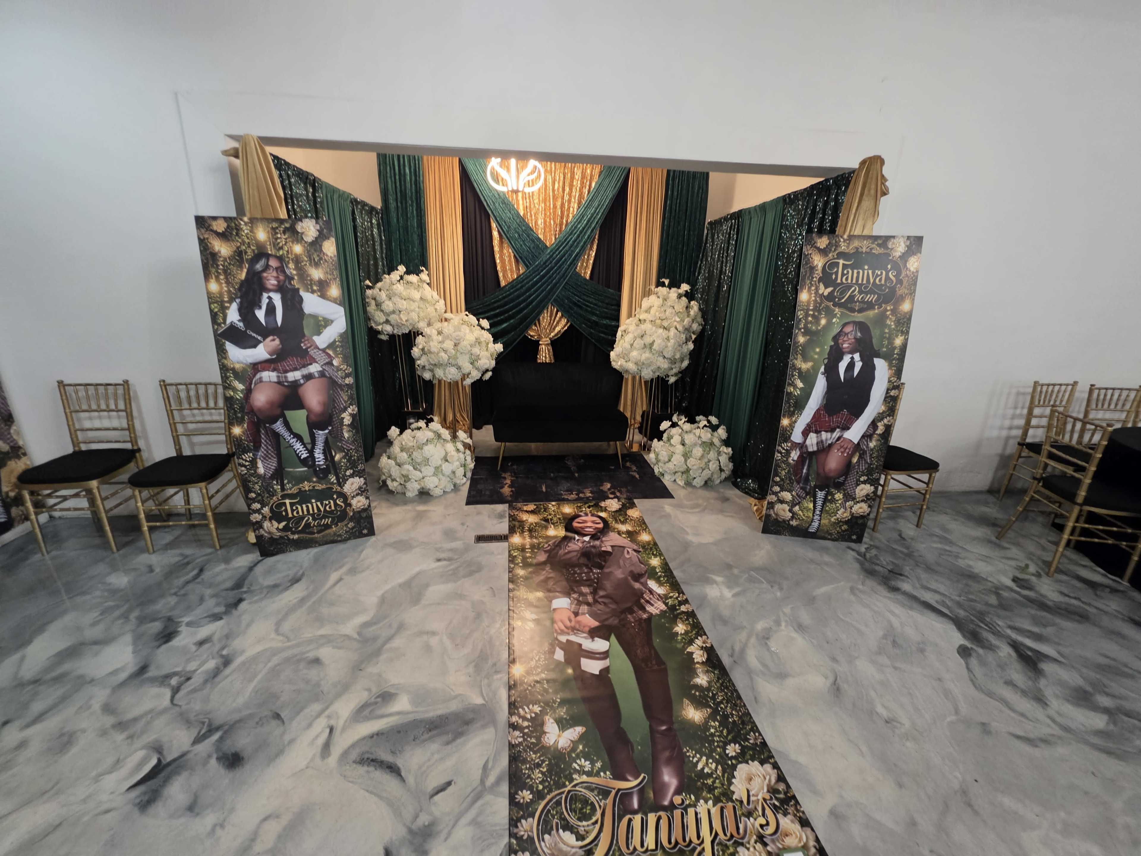 The image shows a decorated event space featuring large banners with a person's image, floral arrangements, and a carpet leading to a central seating area.