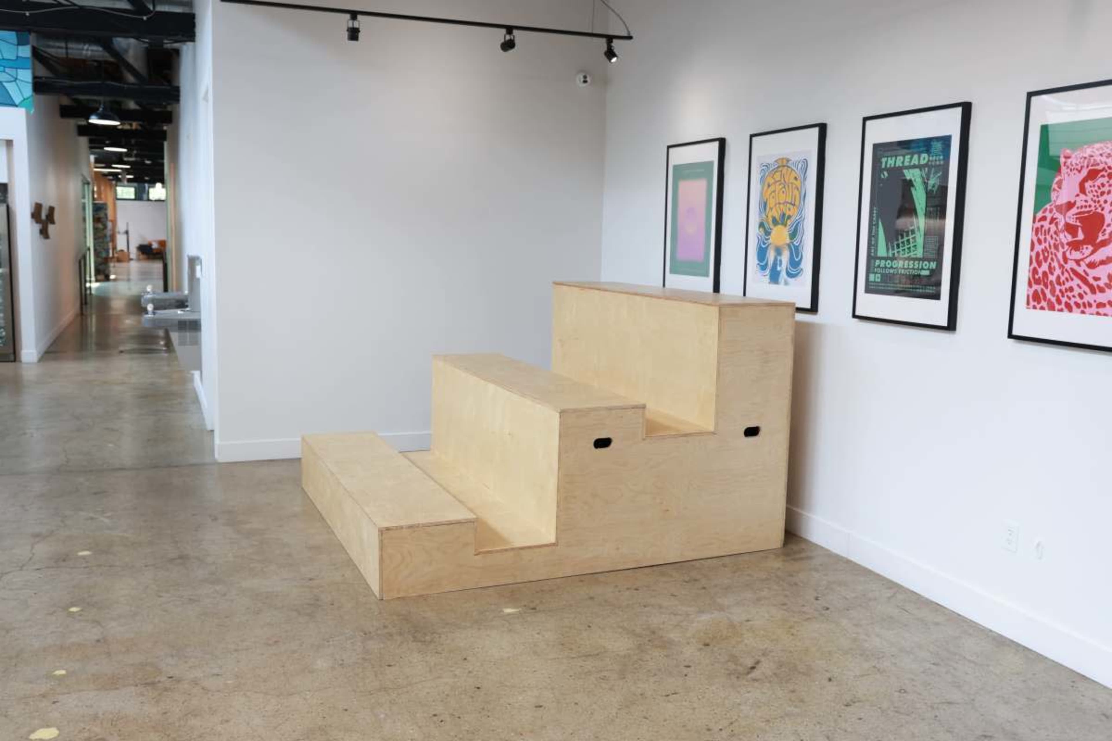 The image shows a three-step wooden structure with a smooth finish positioned against a wall in a modern space, accompanied by framed art on the adjacent wall.
