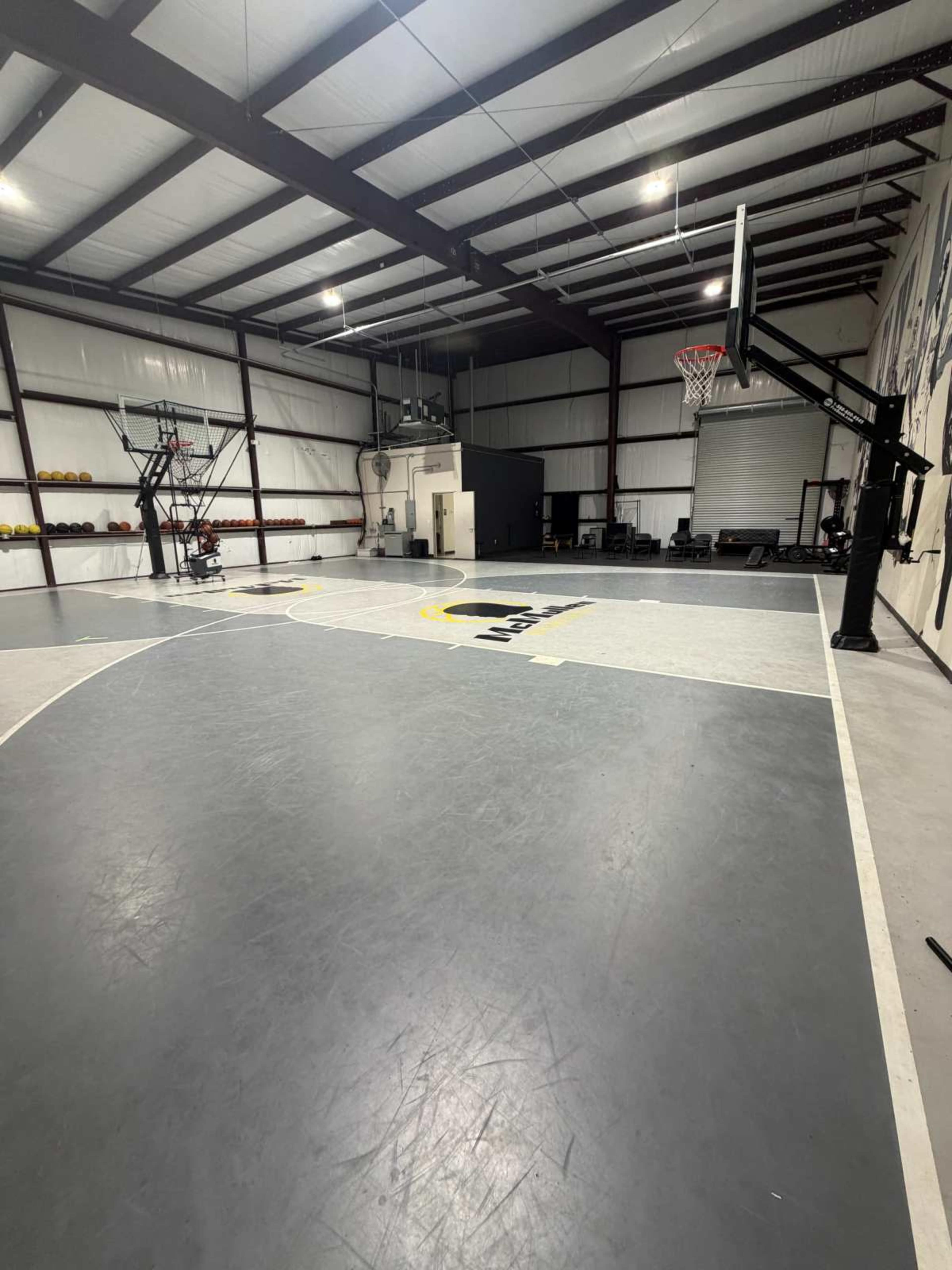 The image shows an indoor basketball court with a hoop, practice equipment, and a clear open space on a gray and white floor.