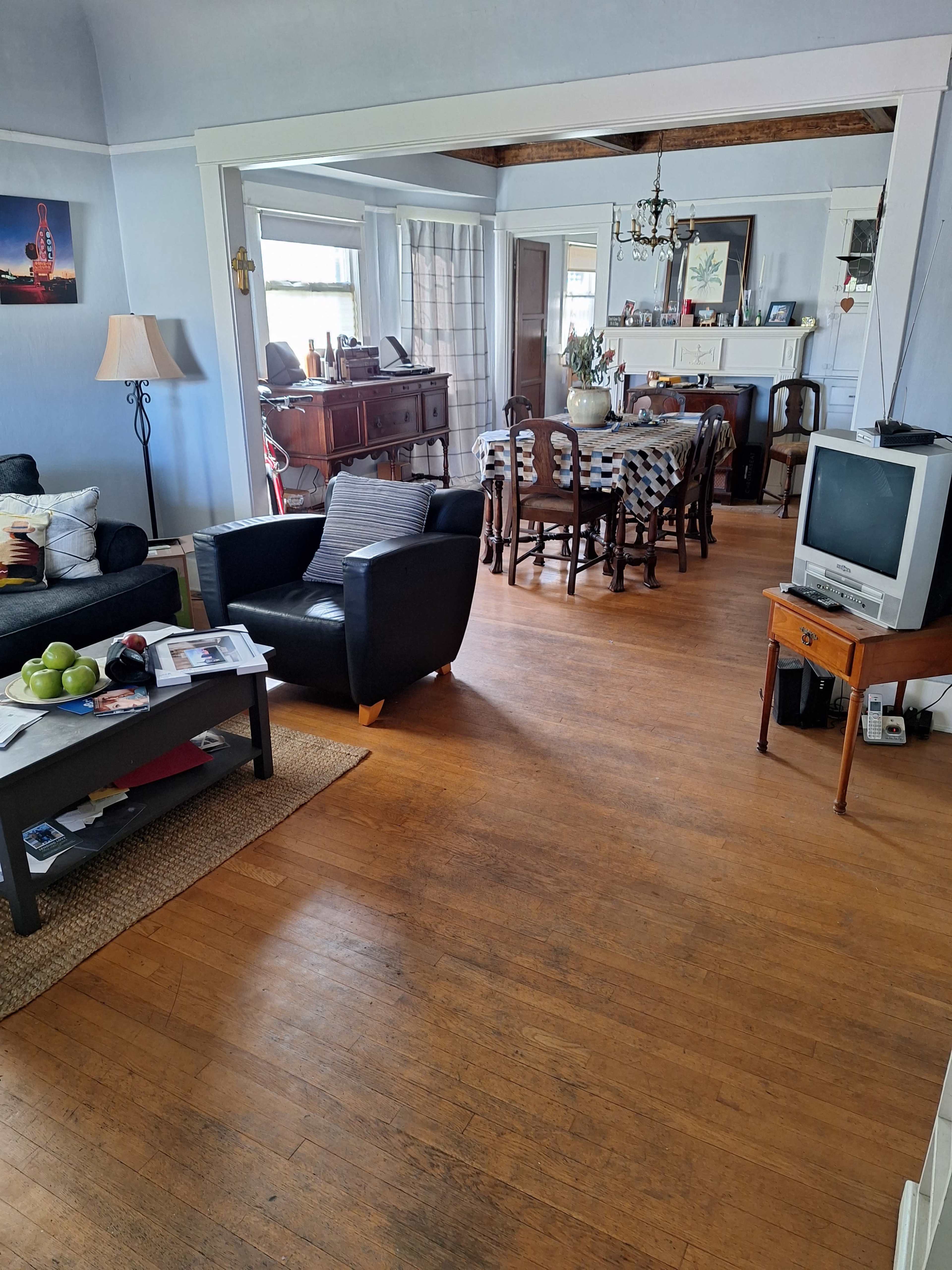 The image shows a spacious living area with a couch and armchair, a dining table set for multiple people, and a television next to a small wooden side table.