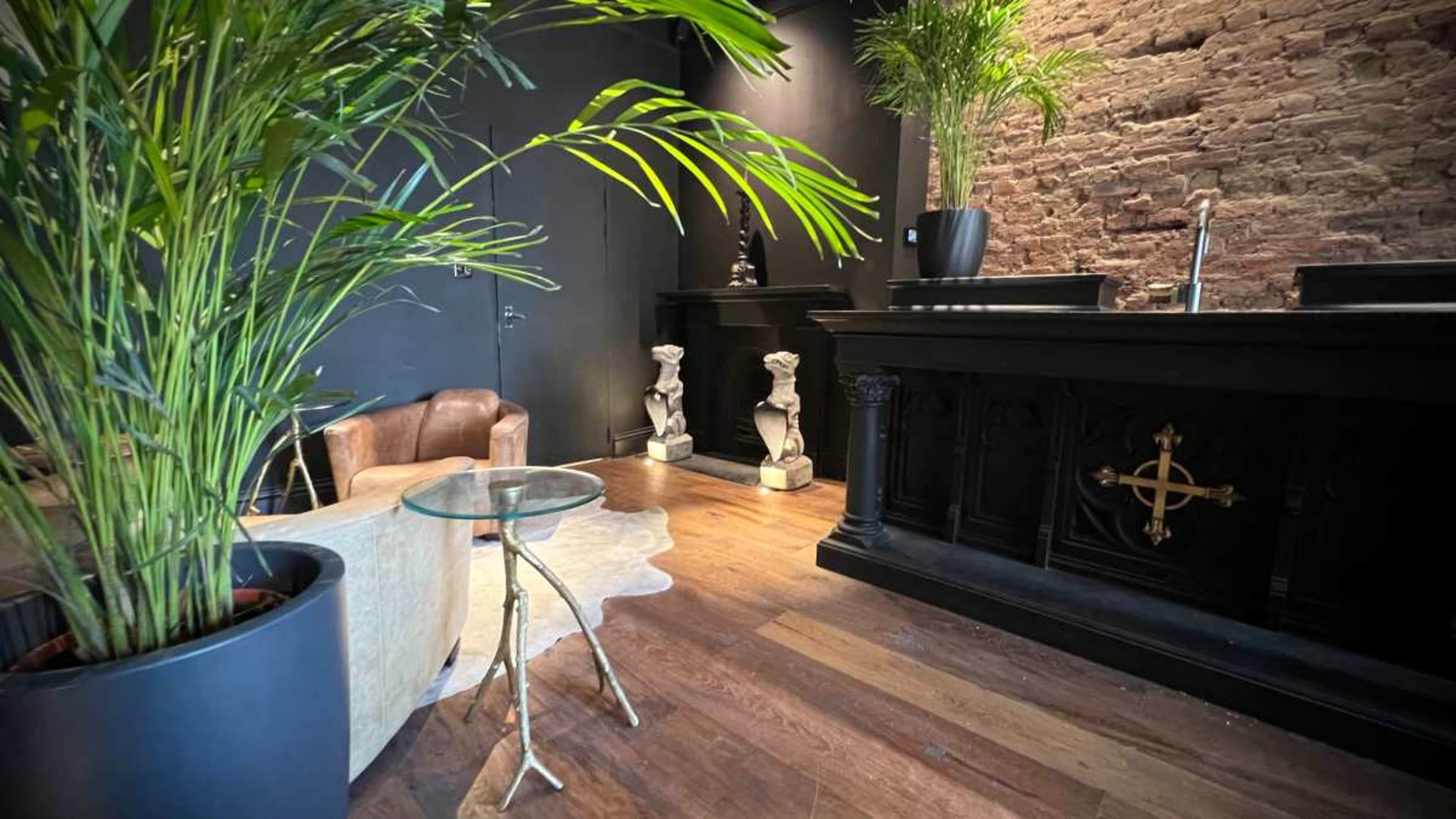 The image shows a modern reception area with a dark wall, wooden flooring, potted plants, a small glass table, a leather armchair, and decorative sculptures.