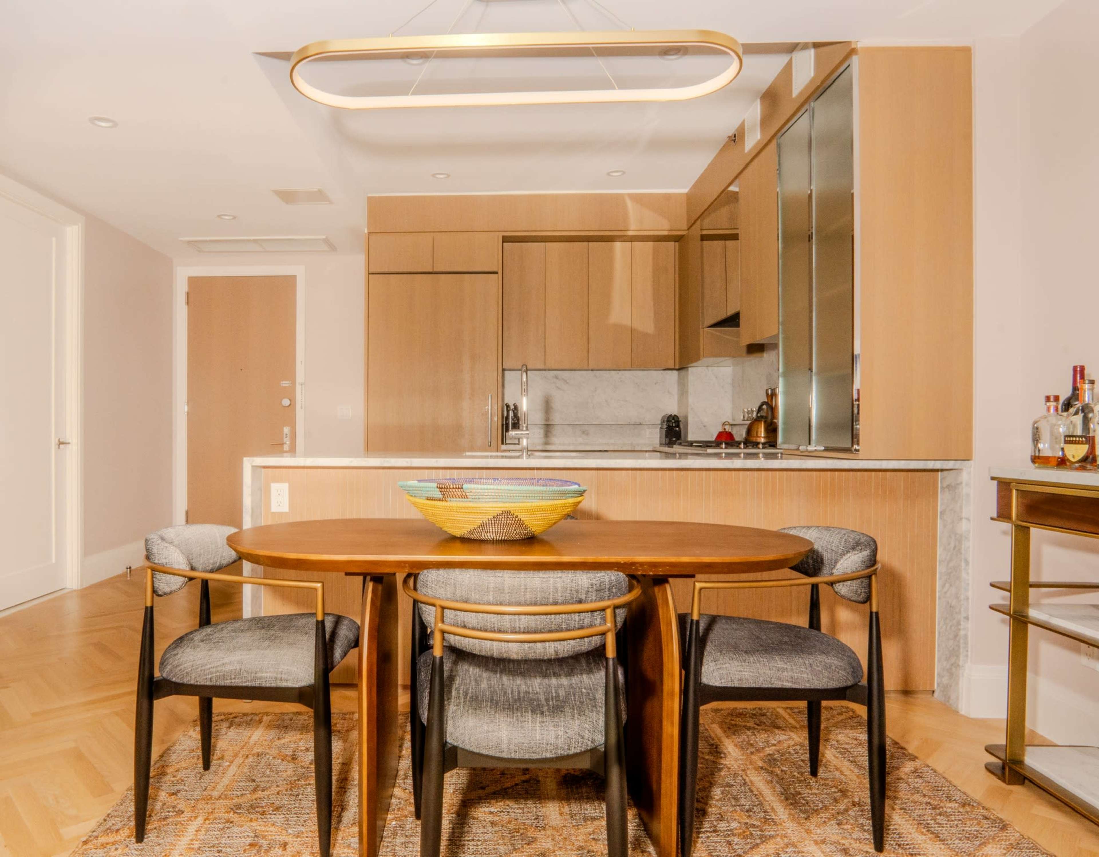 The image shows a modern dining area with a round wooden table and four upholstered chairs, set against a backdrop of a sleek kitchen with wooden cabinetry and a marble countertop.