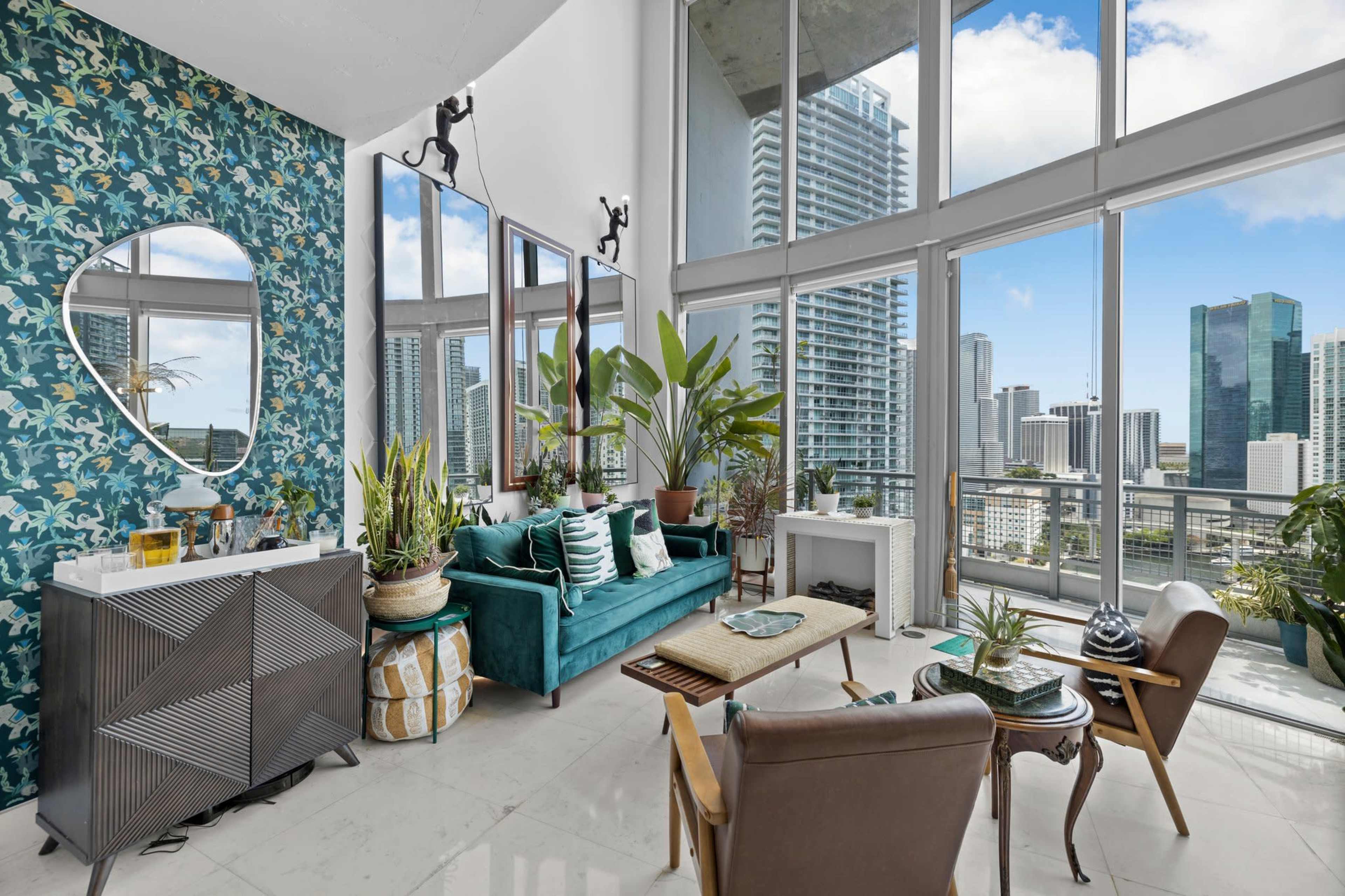A modern living room features large windows with city views, a teal sofa, a mirrored wall, and various indoor plants.