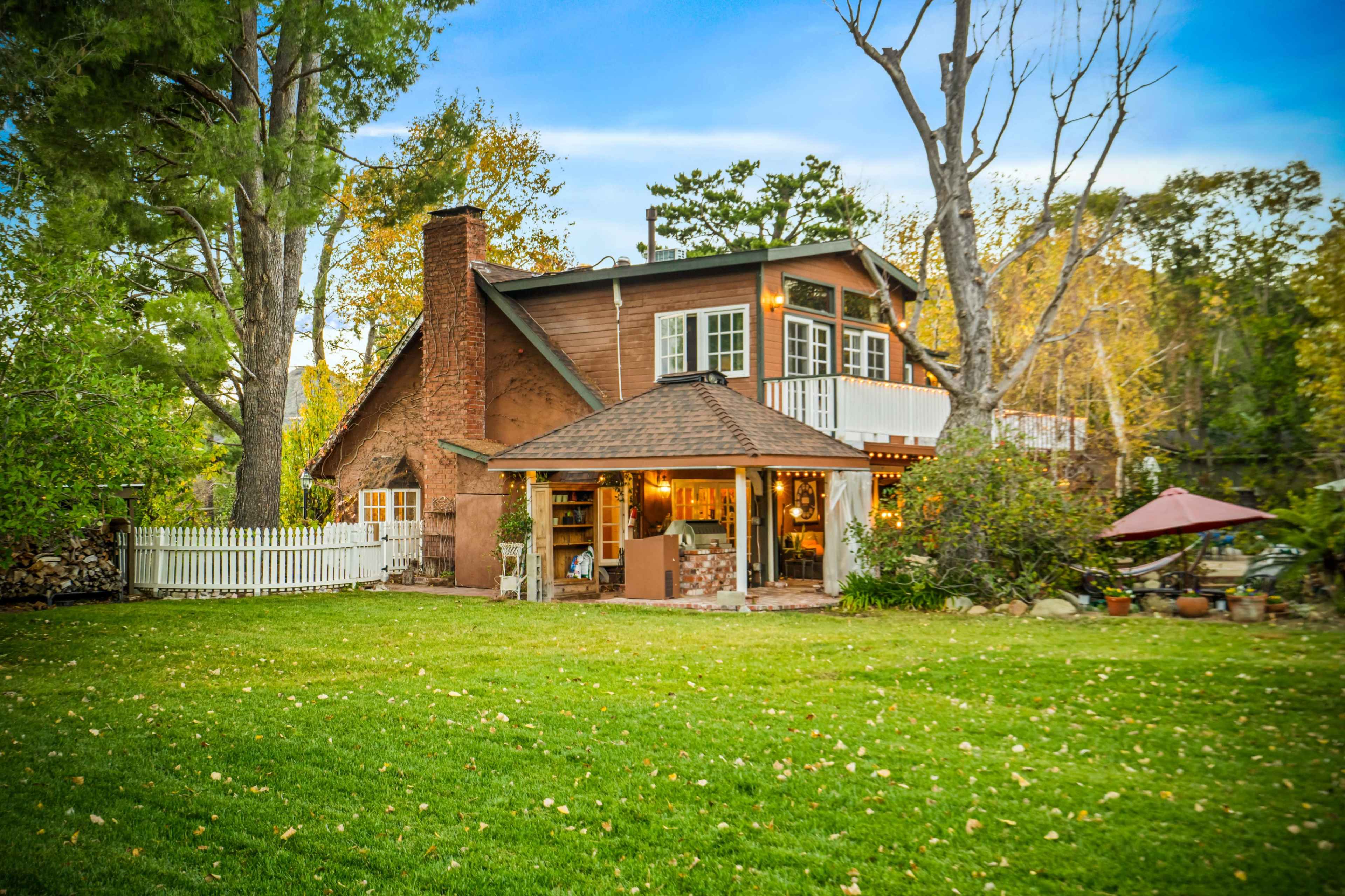 A two-story wooden house with a stone chimney and a covered porch is set amidst a backyard with green grass and trees.