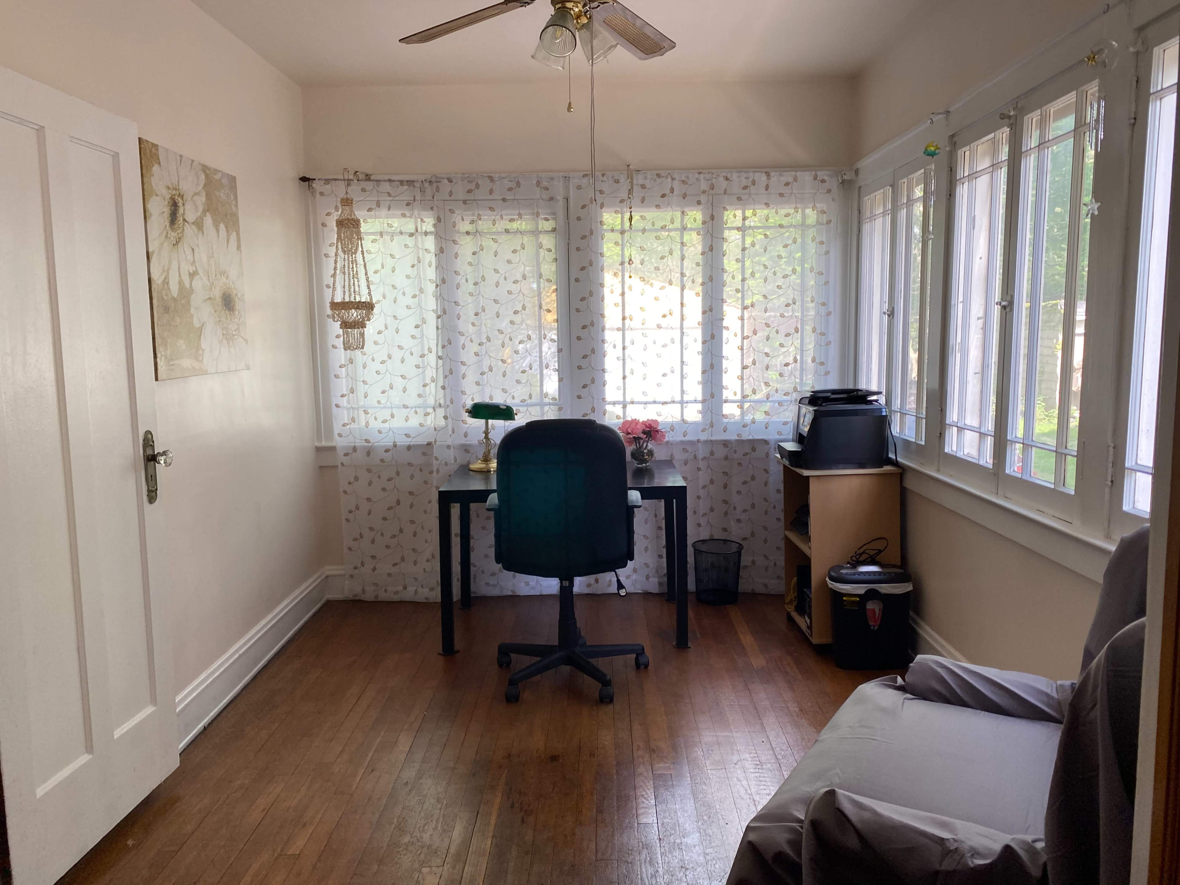 A small office space features a desk and chair near a window with sheer curtains, along with a small shelf and houseplants.