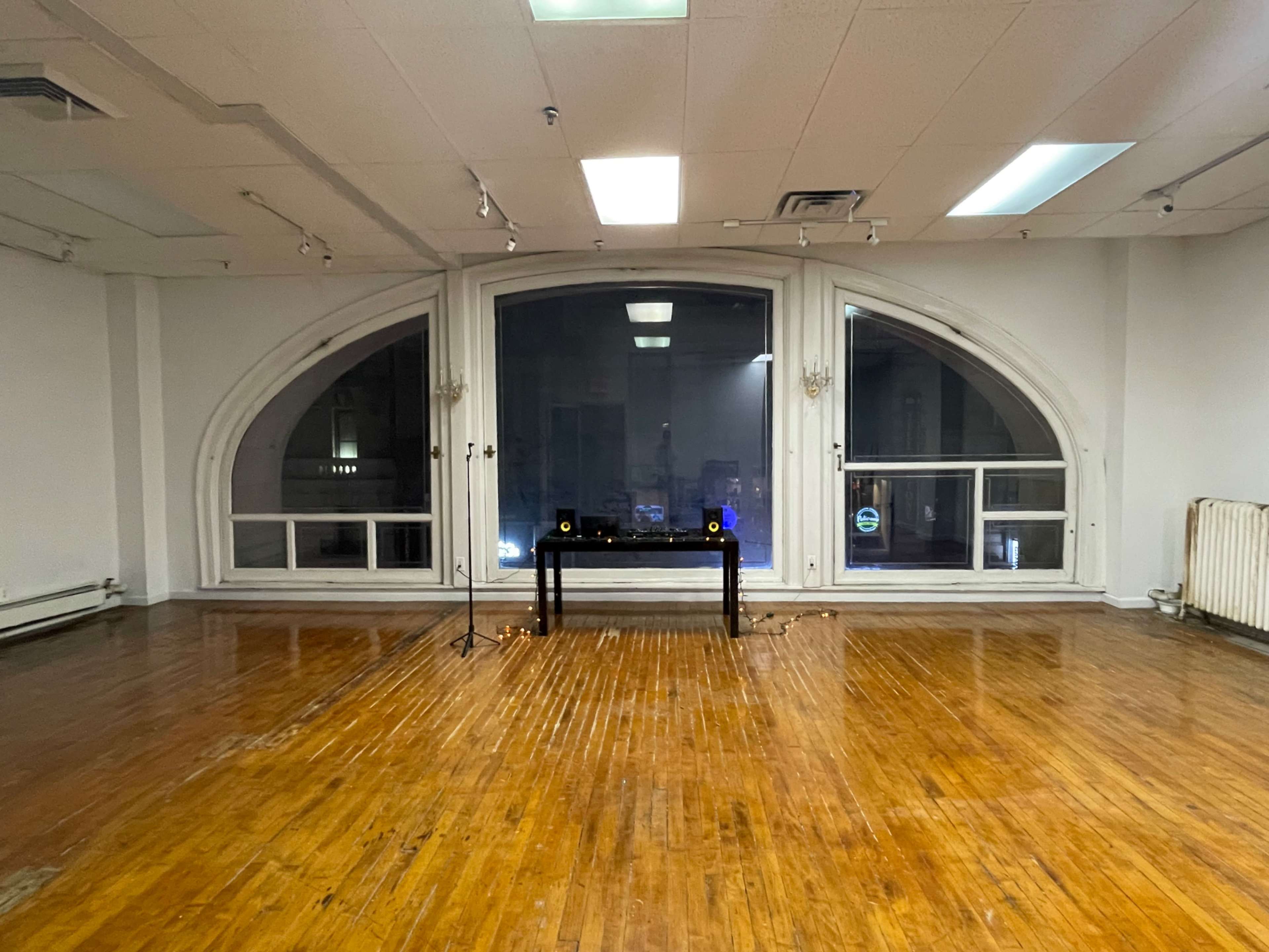 The image shows an empty room with wooden flooring, large arched windows, and a table with speakers positioned in front of the windows.
