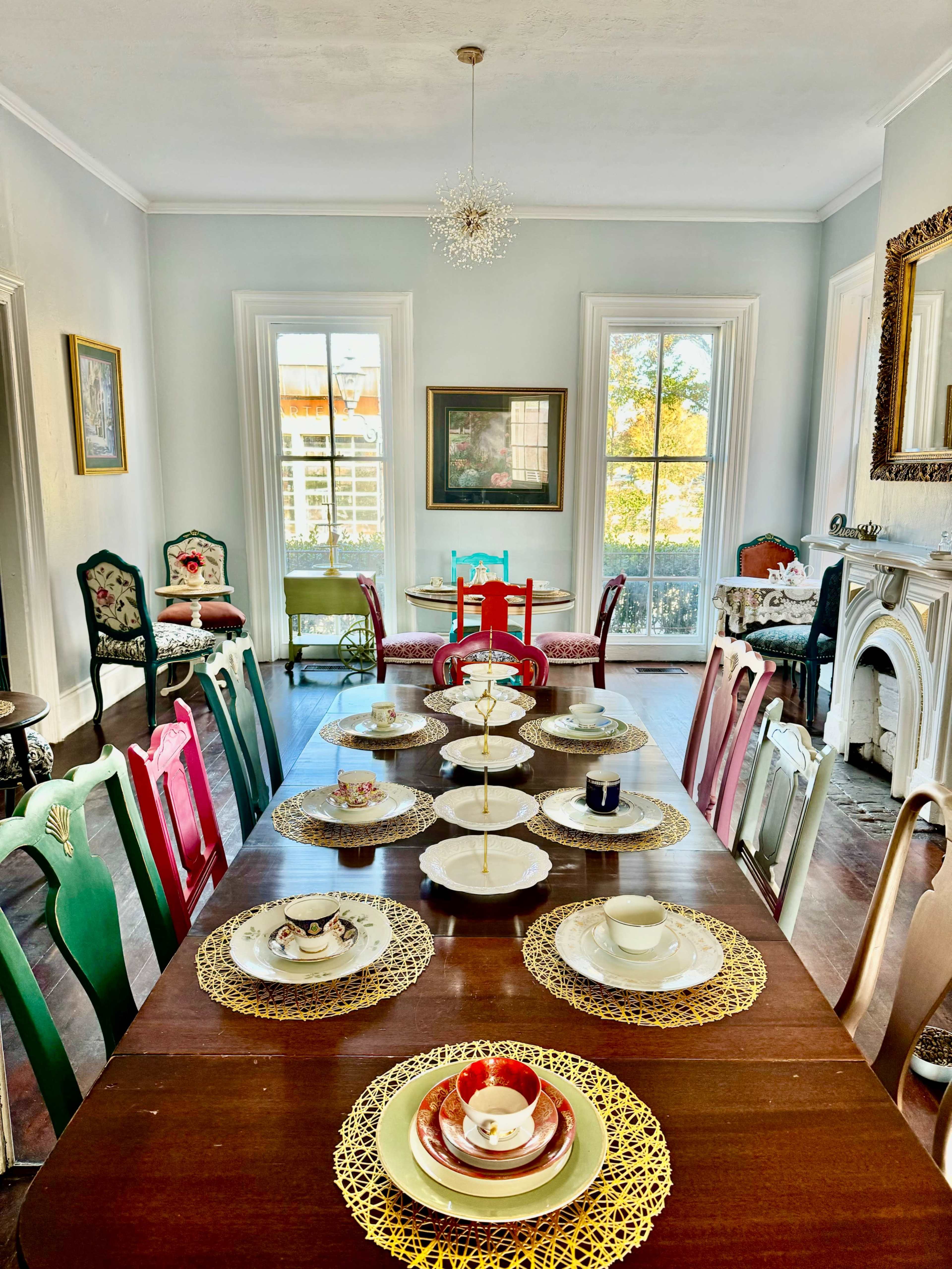 The image shows a dining room with a long wooden table set with plates and decorative items, surrounded by various styled chairs and large windows allowing natural light to fill the space.