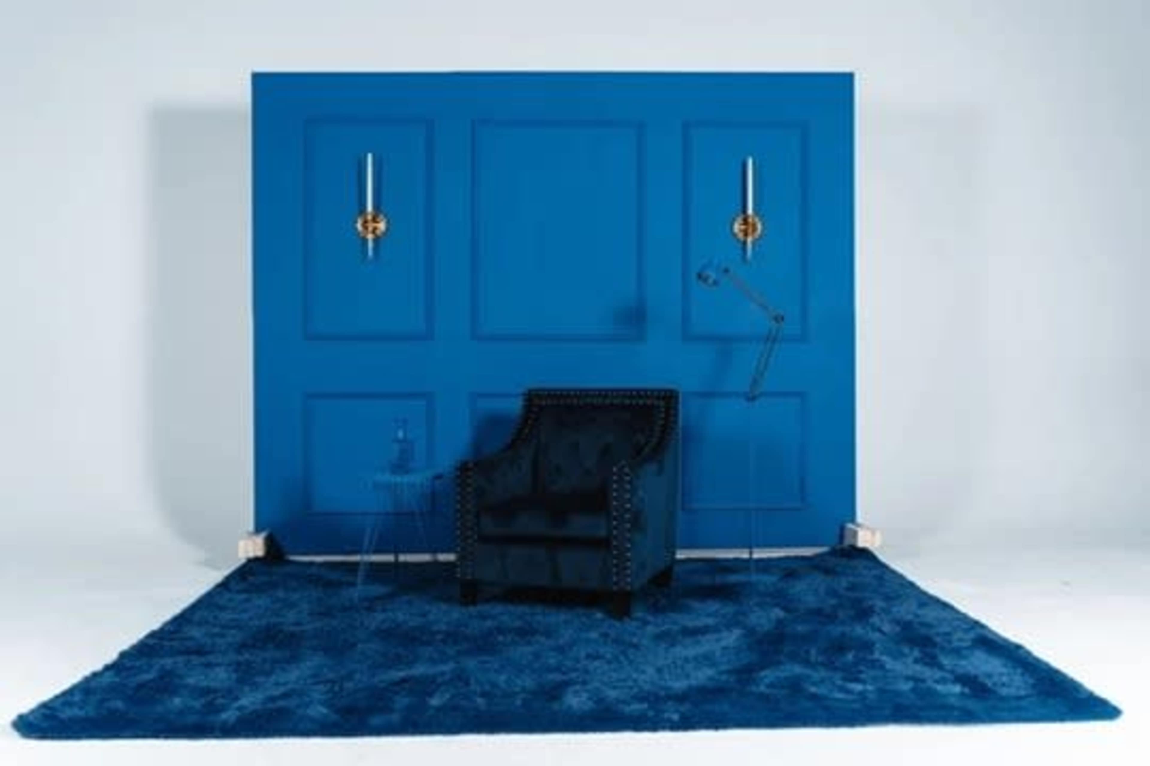 A blue wall with two gold fixtures features a dark velvet armchair in front of a clear side table and a floor covered with a shaggy blue rug.
