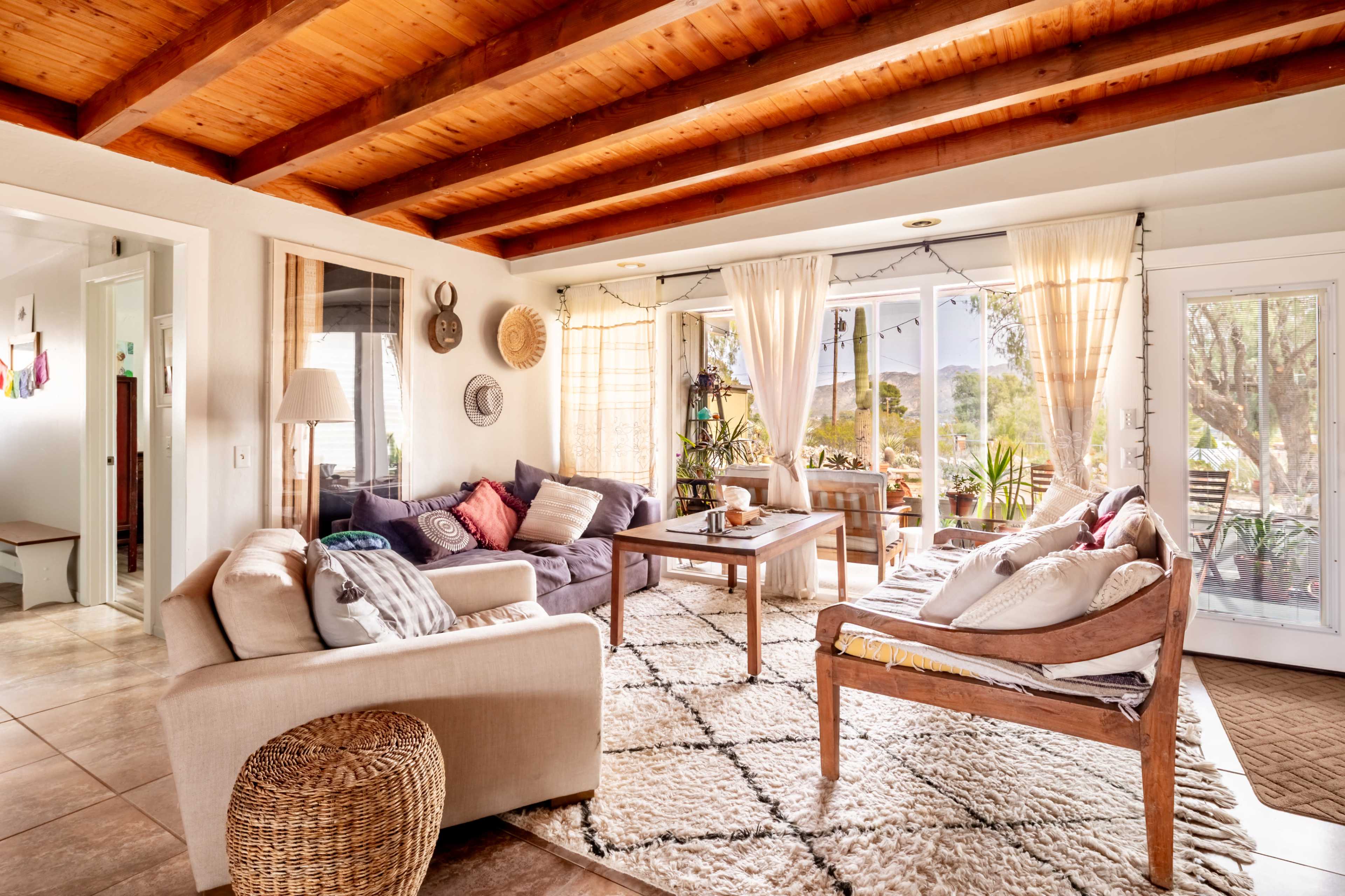 The image shows a bright, open living room with wooden beams, large windows, a mix of seating options, and plants, creating a cozy atmosphere.