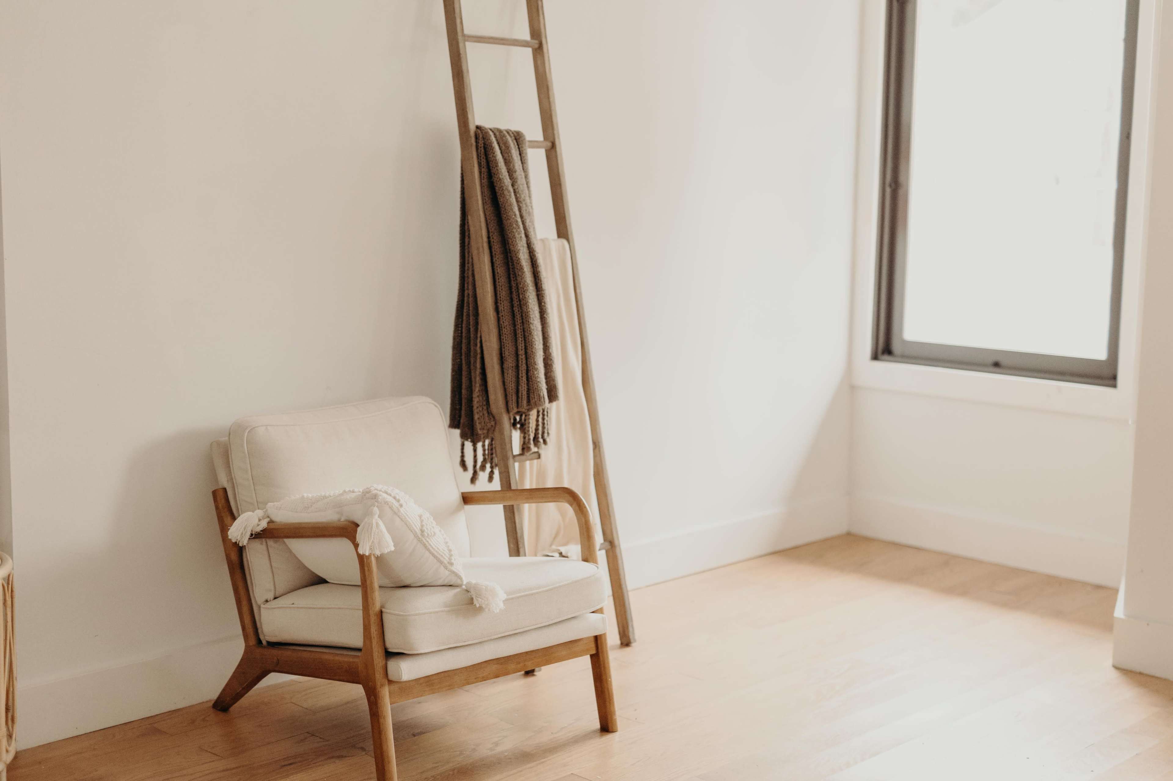 A wooden ladder holding folded blankets leans against a white wall near a beige armchair in a sparsely furnished room with a large window.