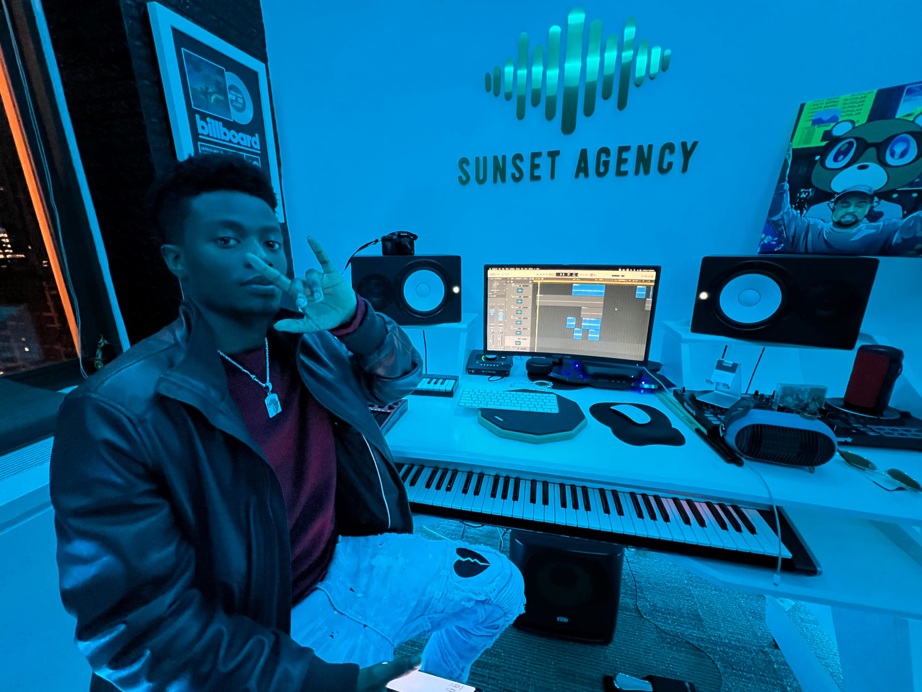 A person sits at a desk in a music studio, surrounded by computer monitors, speakers, and musical equipment, with blue lighting illuminating the space.
