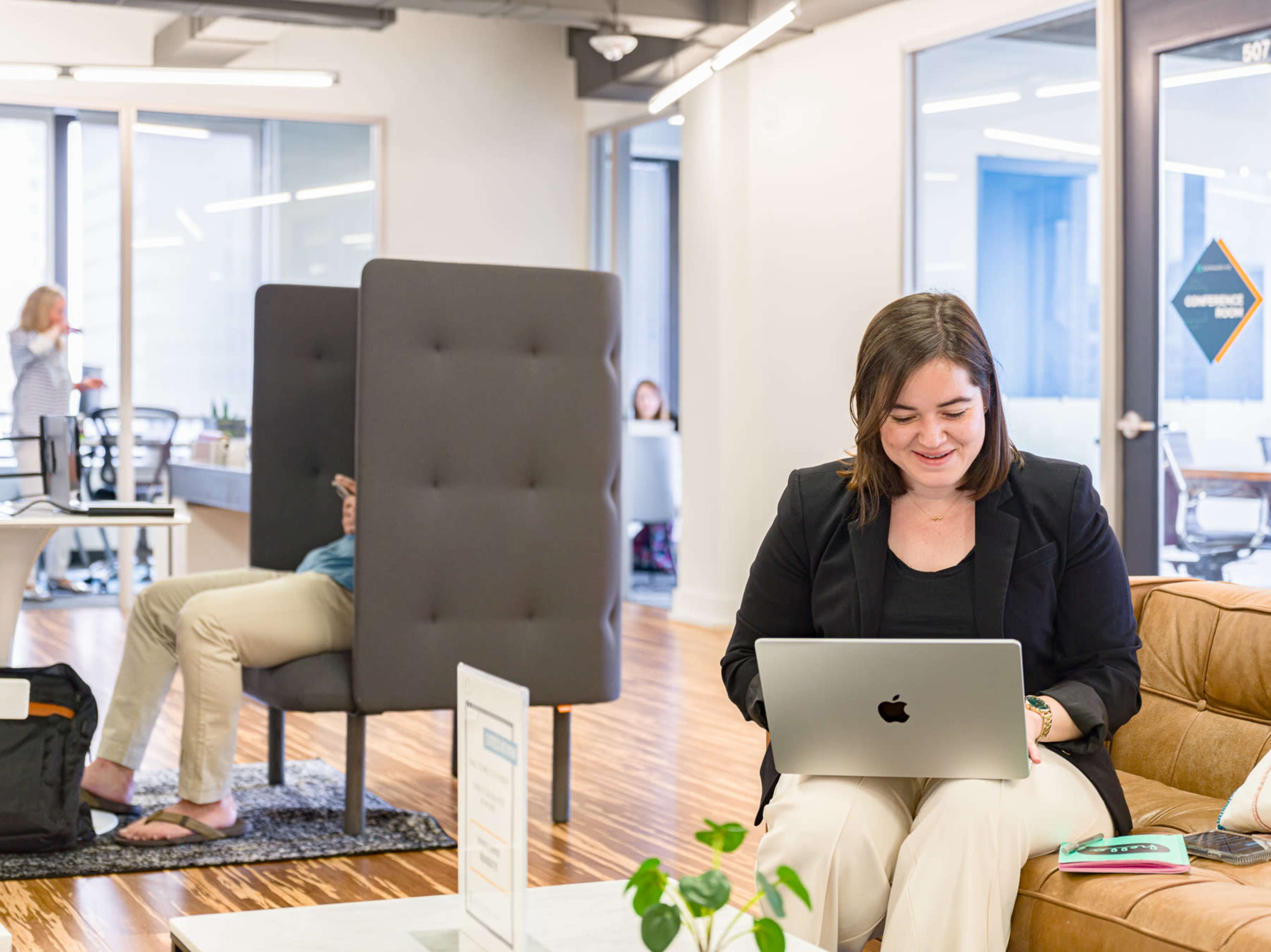 A woman sits on a sofa, working on a laptop in a modern office space, while another person is visible in a seated position behind a privacy screen.