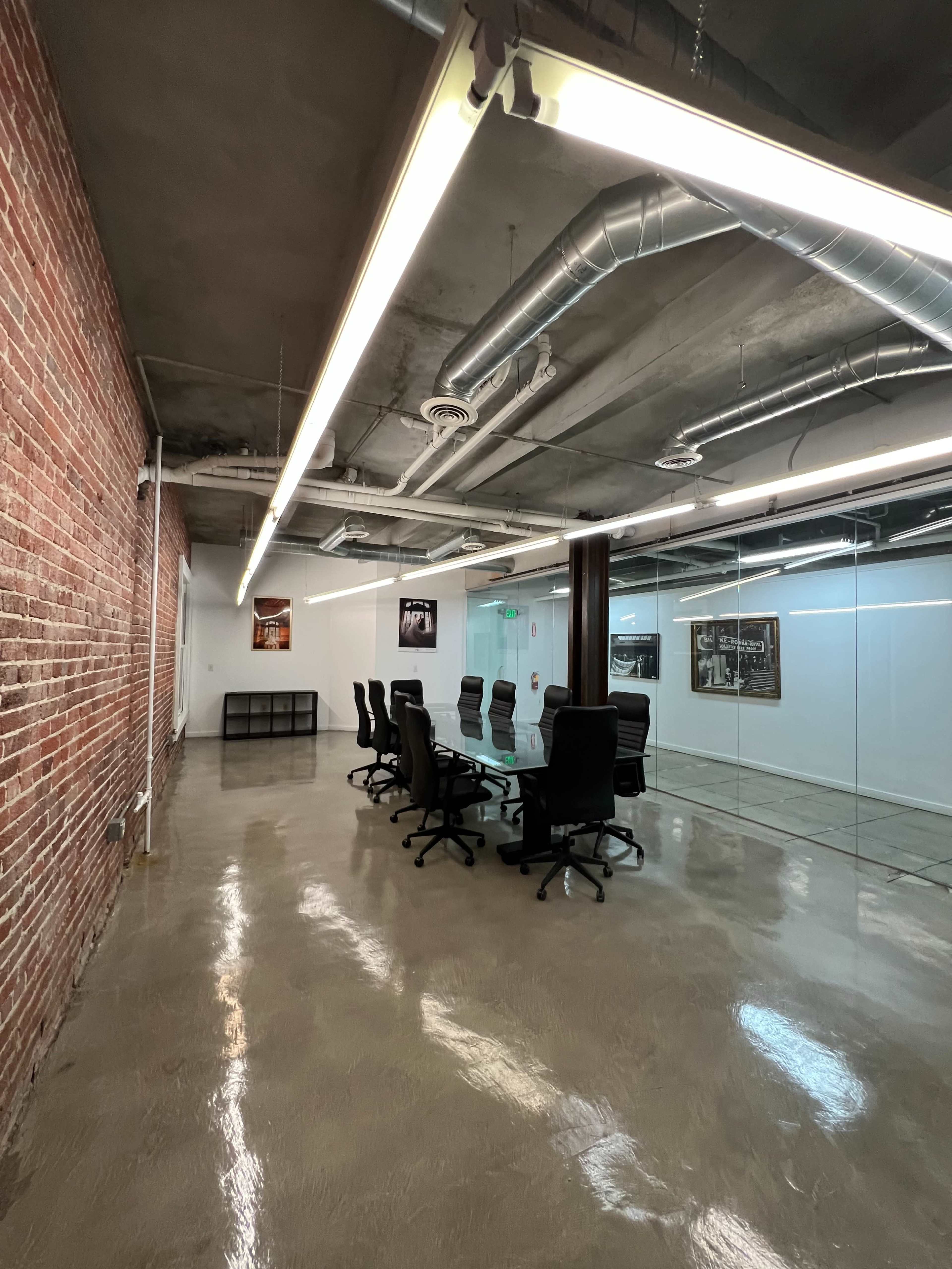 A modern conference room with a glass wall, black office chairs around a long table, and exposed brick on one side.