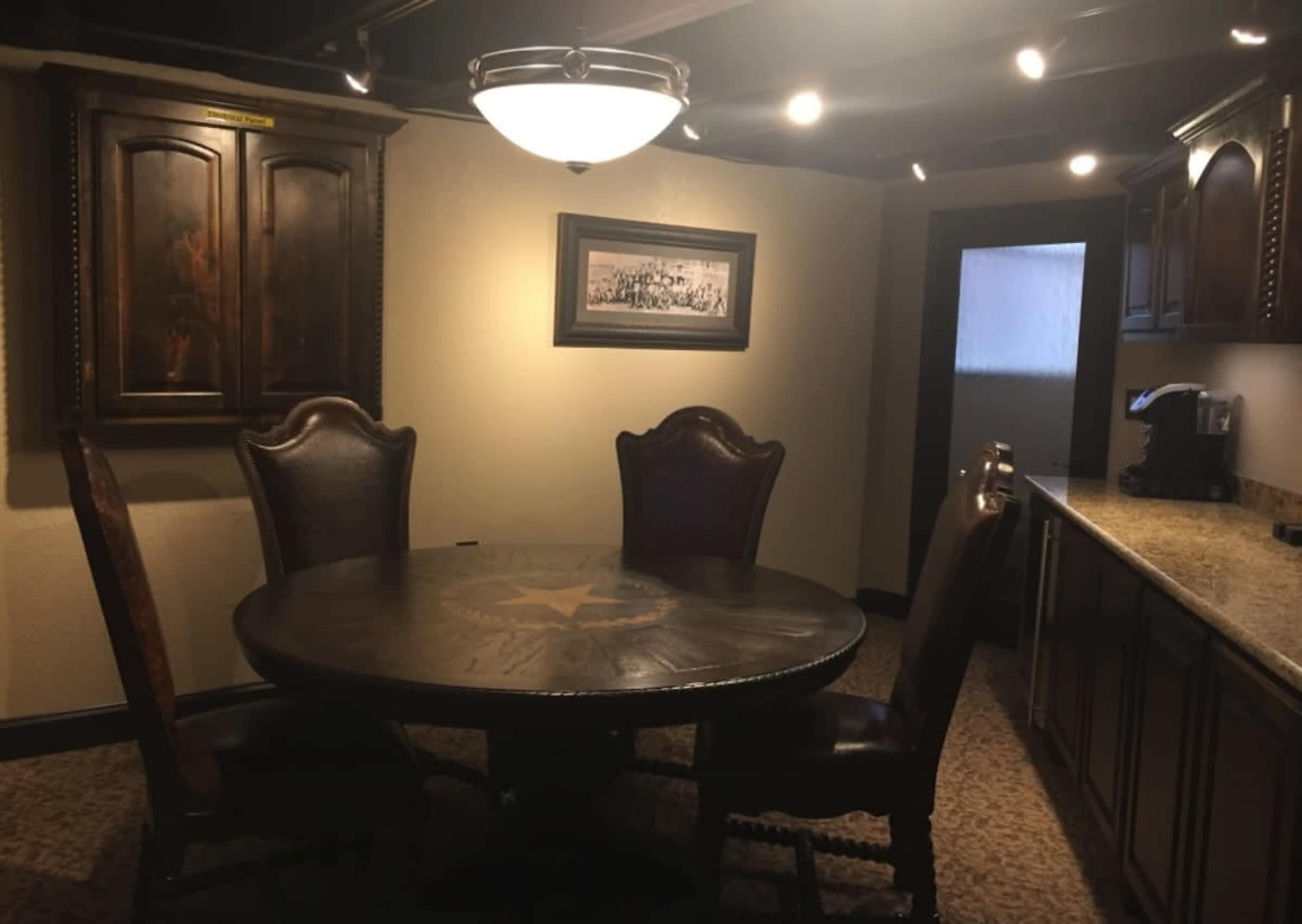 The image shows a dining area with a round wooden table and four high-backed chairs, surrounded by cabinets and a coffee station in a well-lit room.