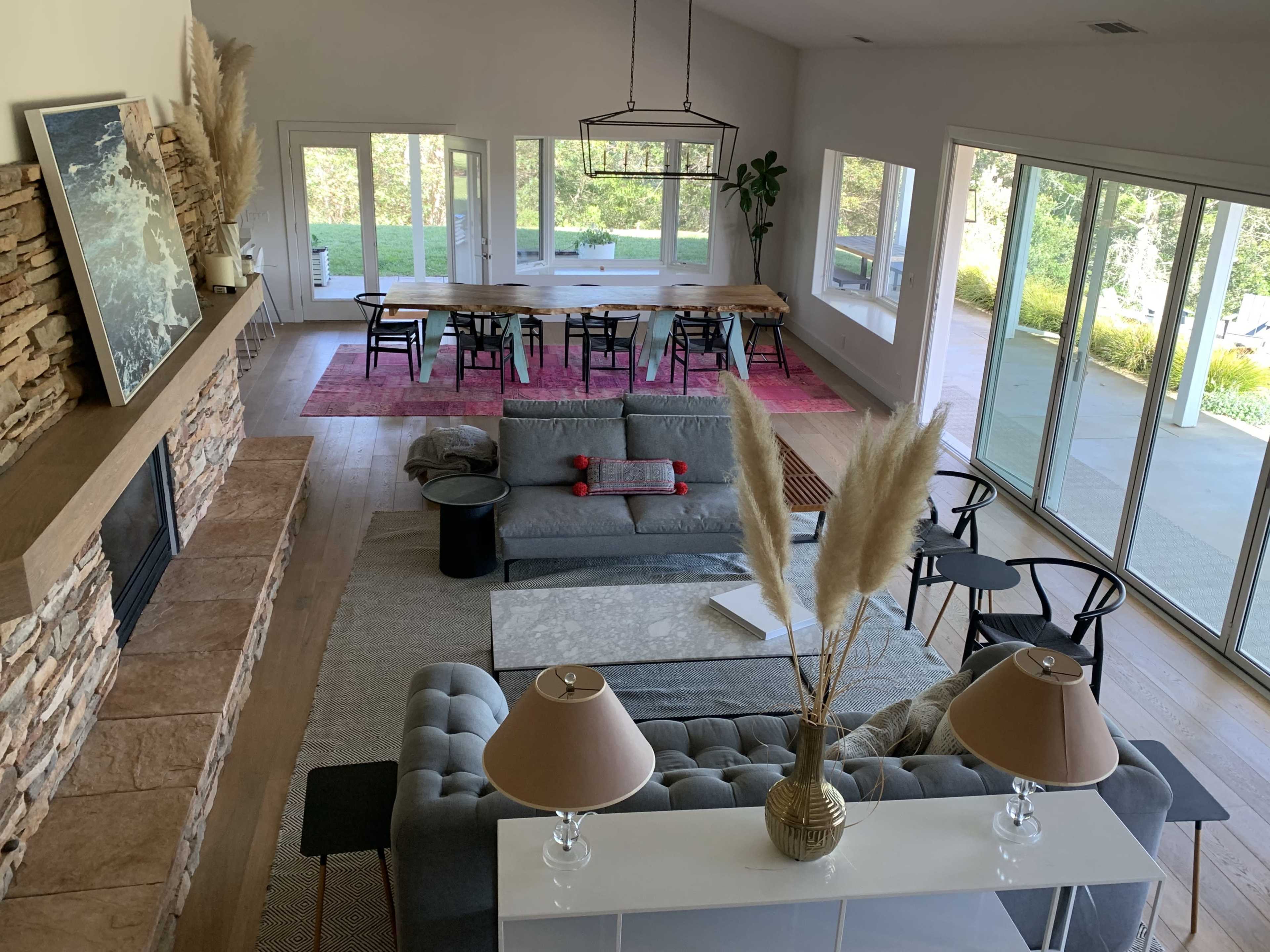 A spacious living room features a gray sofa, a coffee table, and a stone fireplace, with large windows overlooking a dining area and outdoor landscape.