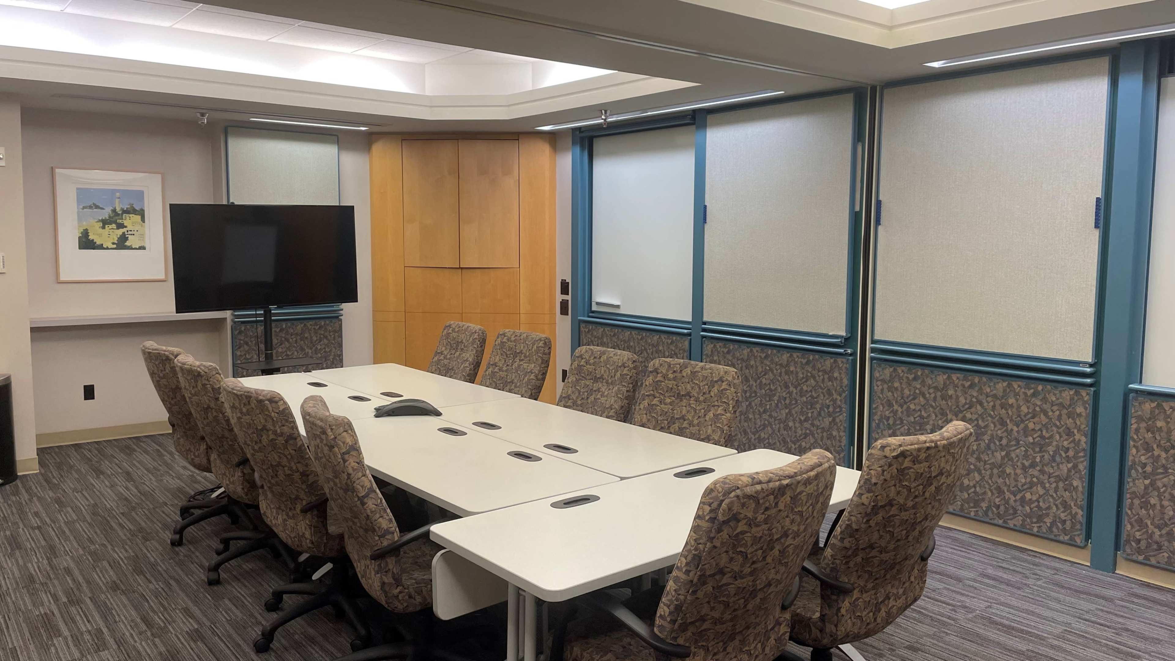 A conference room with a long table, eight chairs, a television screen, and large windows covered by blinds.