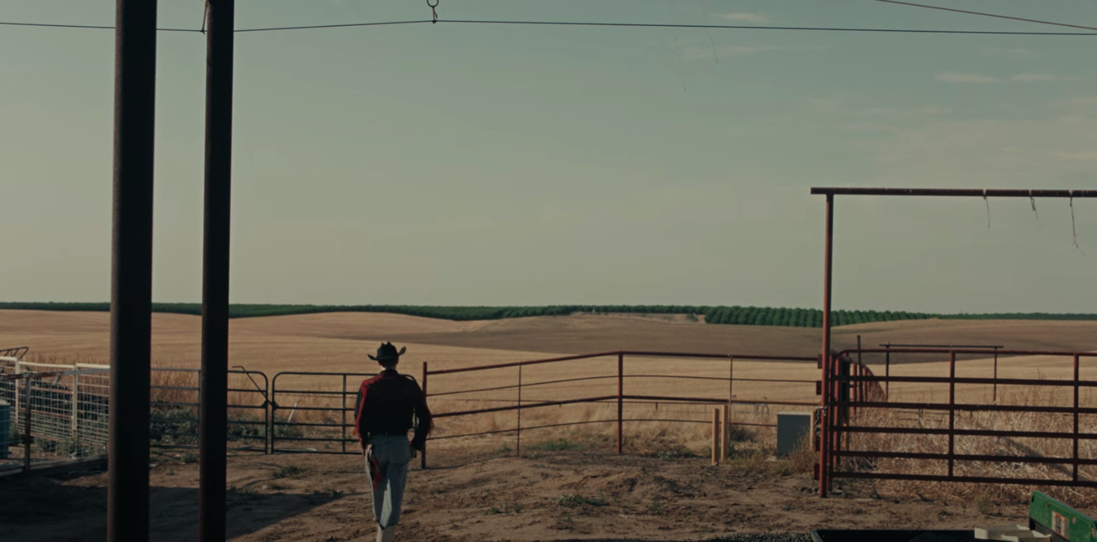 A person in a cowboy hat walks towards an open gate, with fields stretching out in the background.