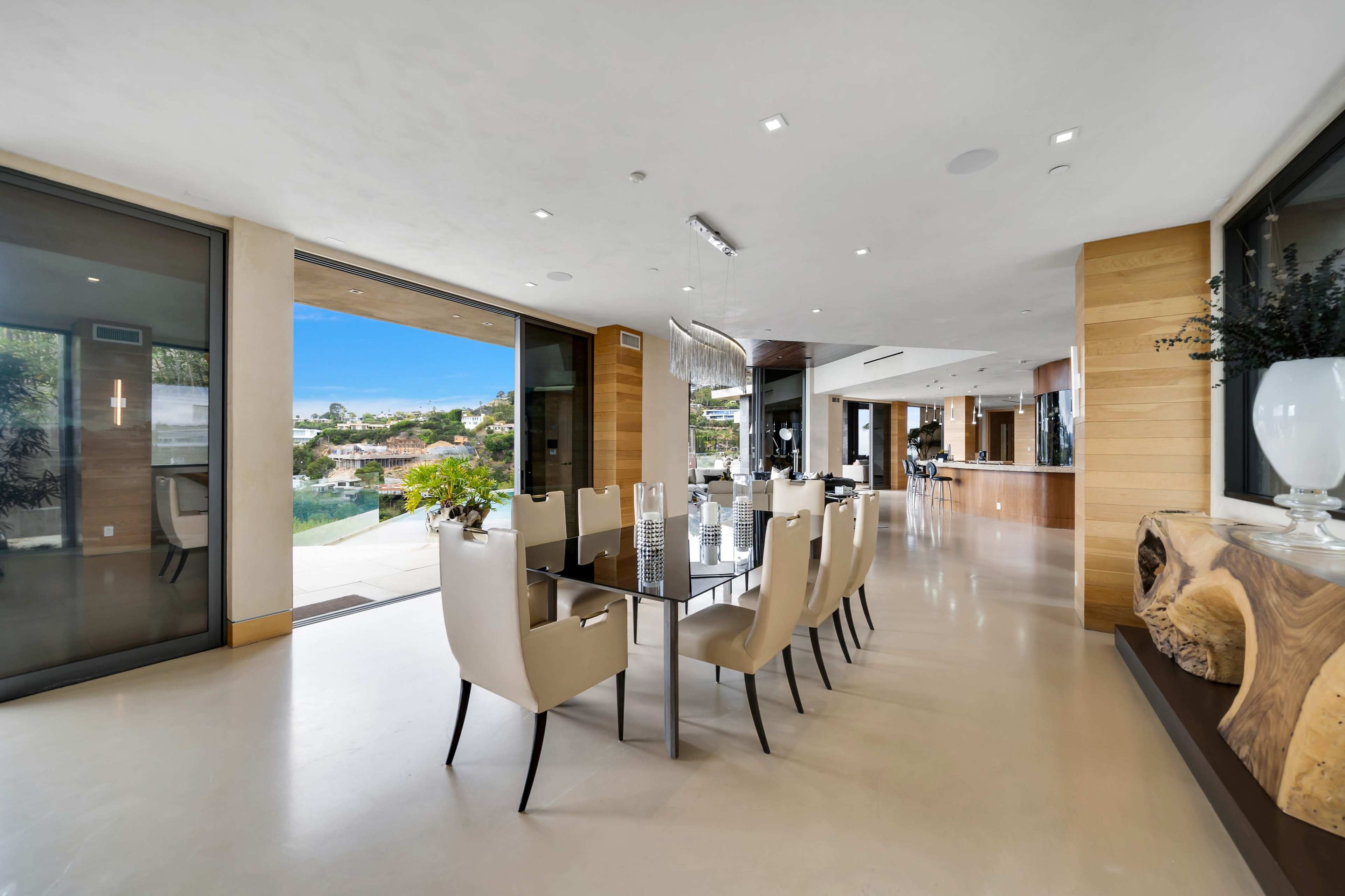 A modern dining area features a glass table surrounded by beige chairs, with large windows showcasing a view of the outdoor landscape.