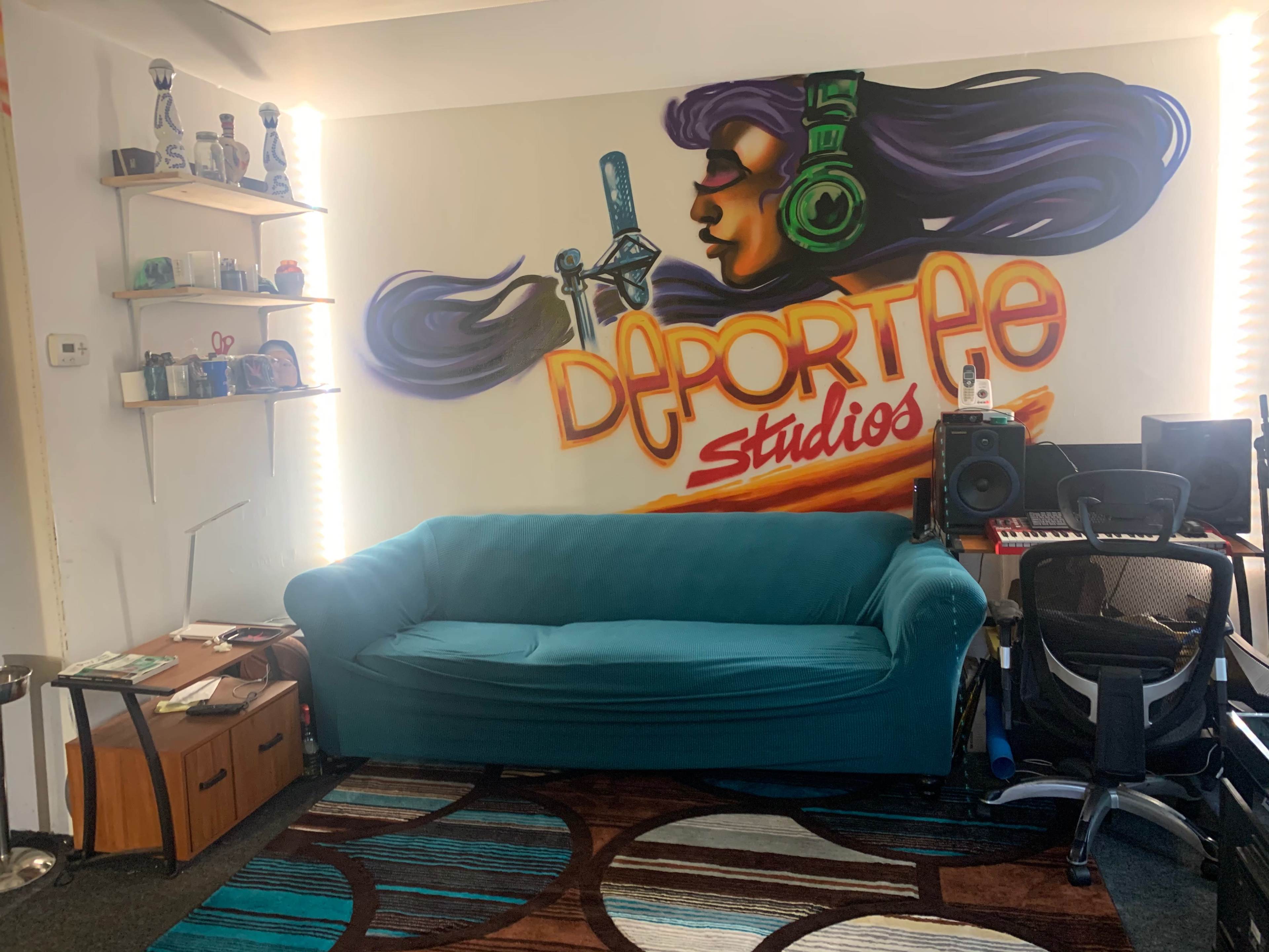 A cozy studio space featuring a turquoise couch, a wall mural with the text "Deportee Studios," and musical equipment arranged in the corner.