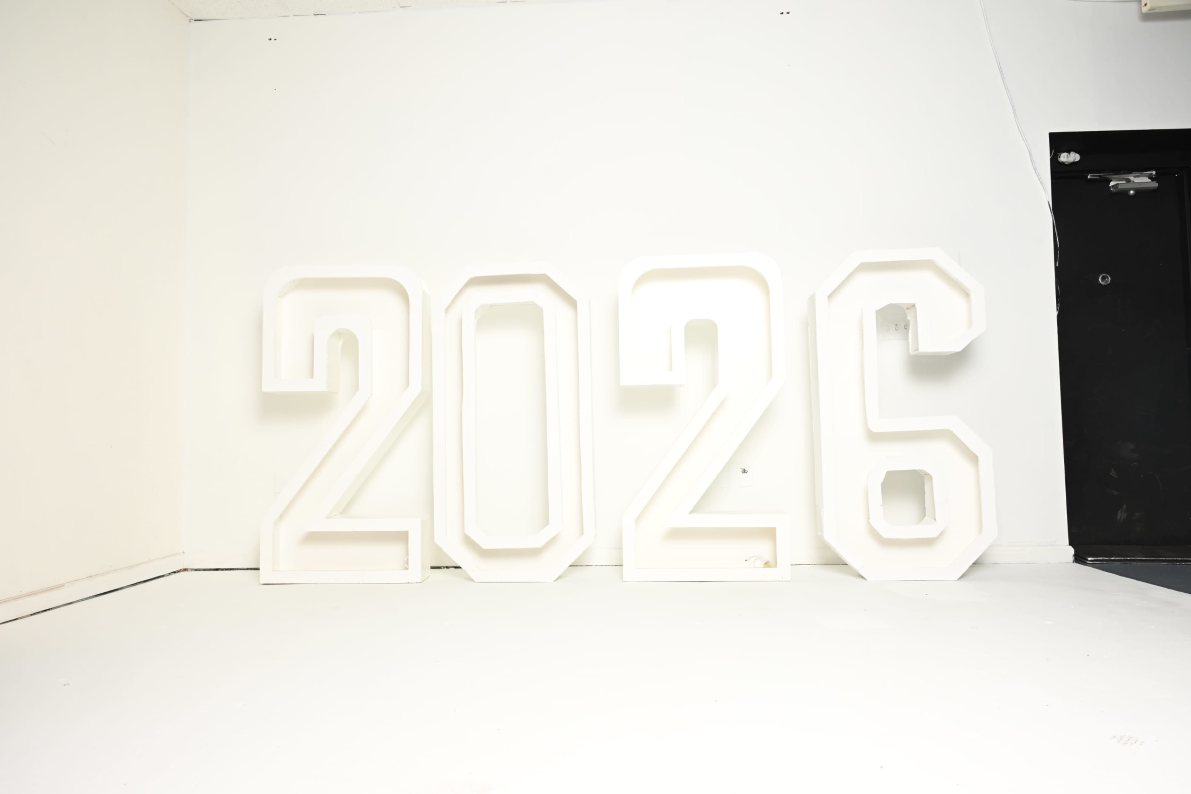 The image shows large, white illuminated numbers "2026" arranged against a plain white wall.
