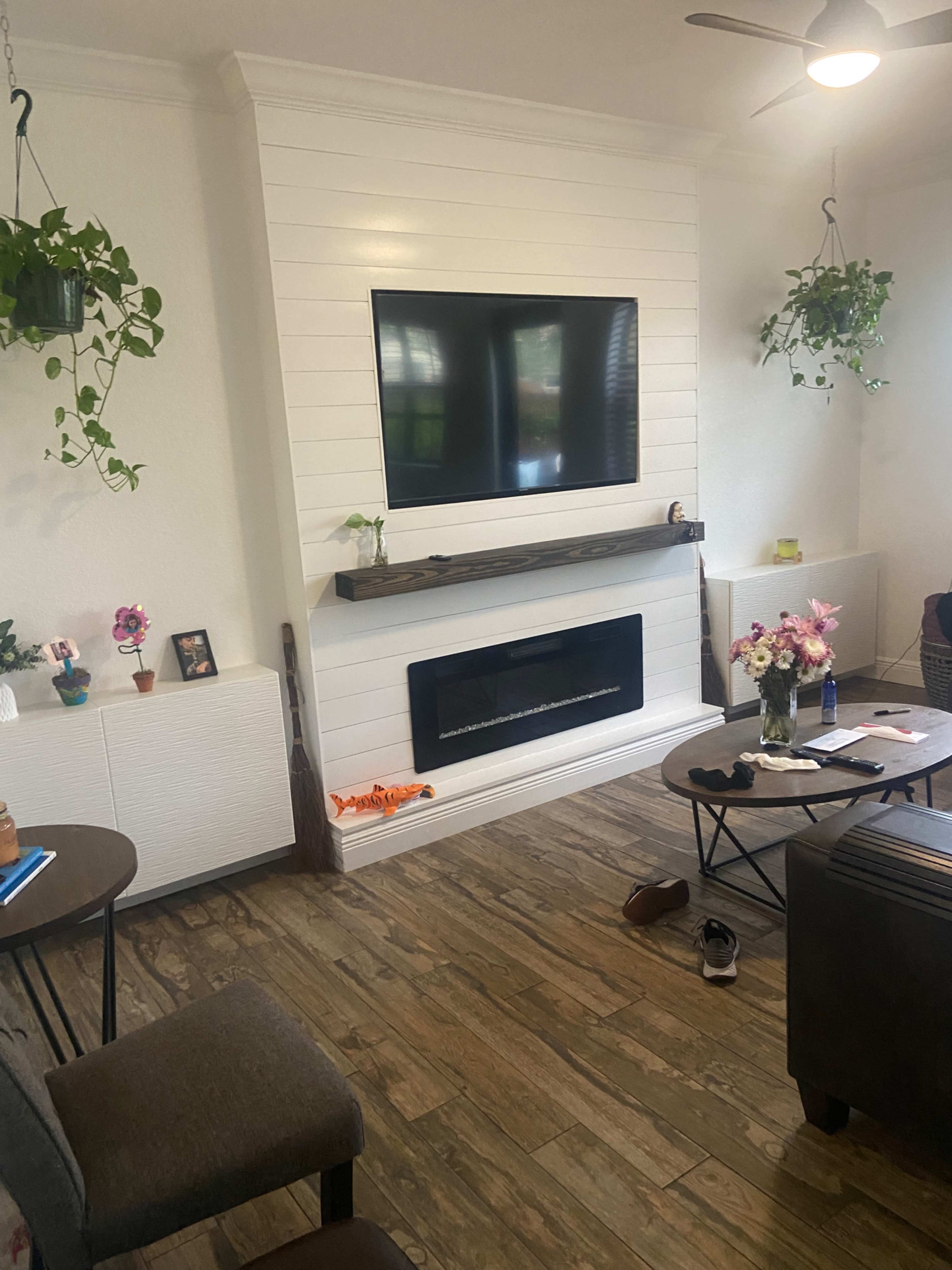 The living room features a white shiplap accent wall with a mounted television and a modern electric fireplace, surrounded by potted plants and a coffee table.