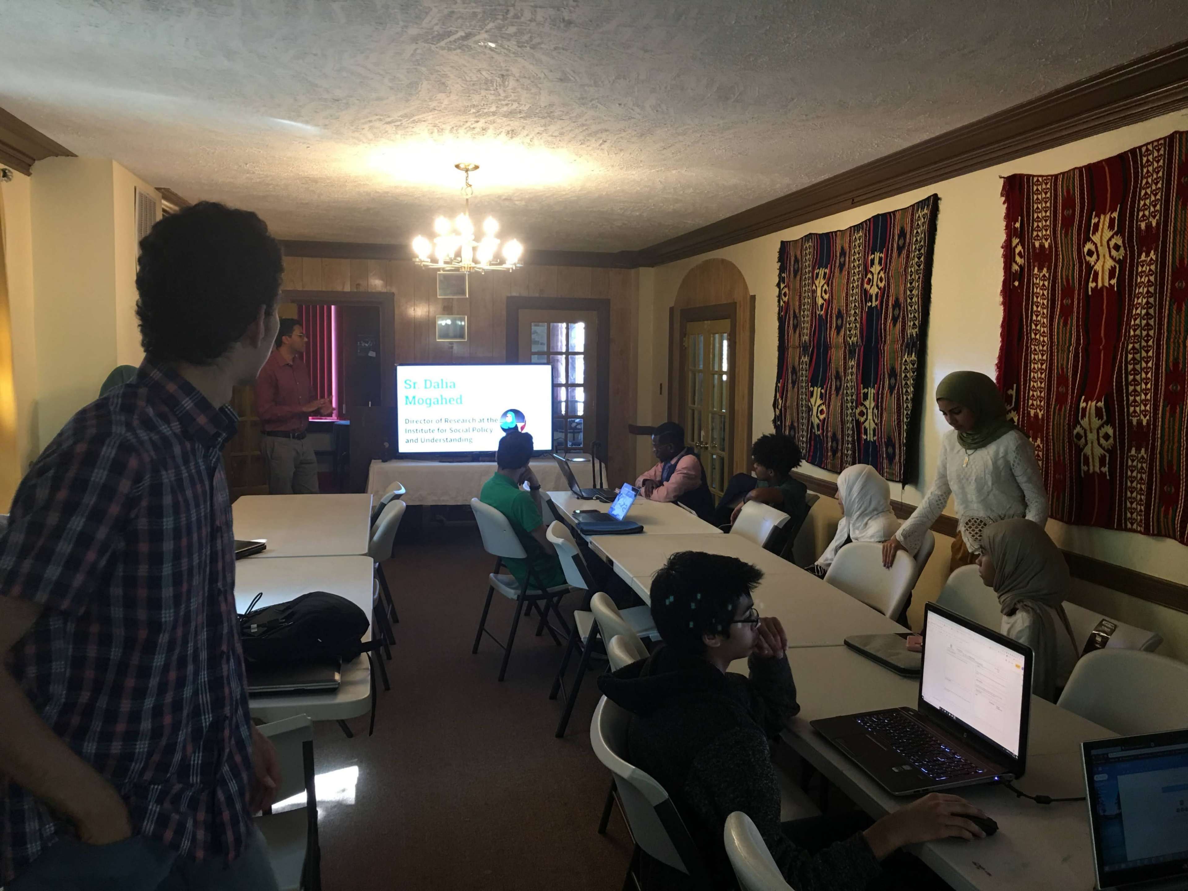 A group of individuals, some seated at tables with laptops, is attending a presentation in a room decorated with traditional textiles.