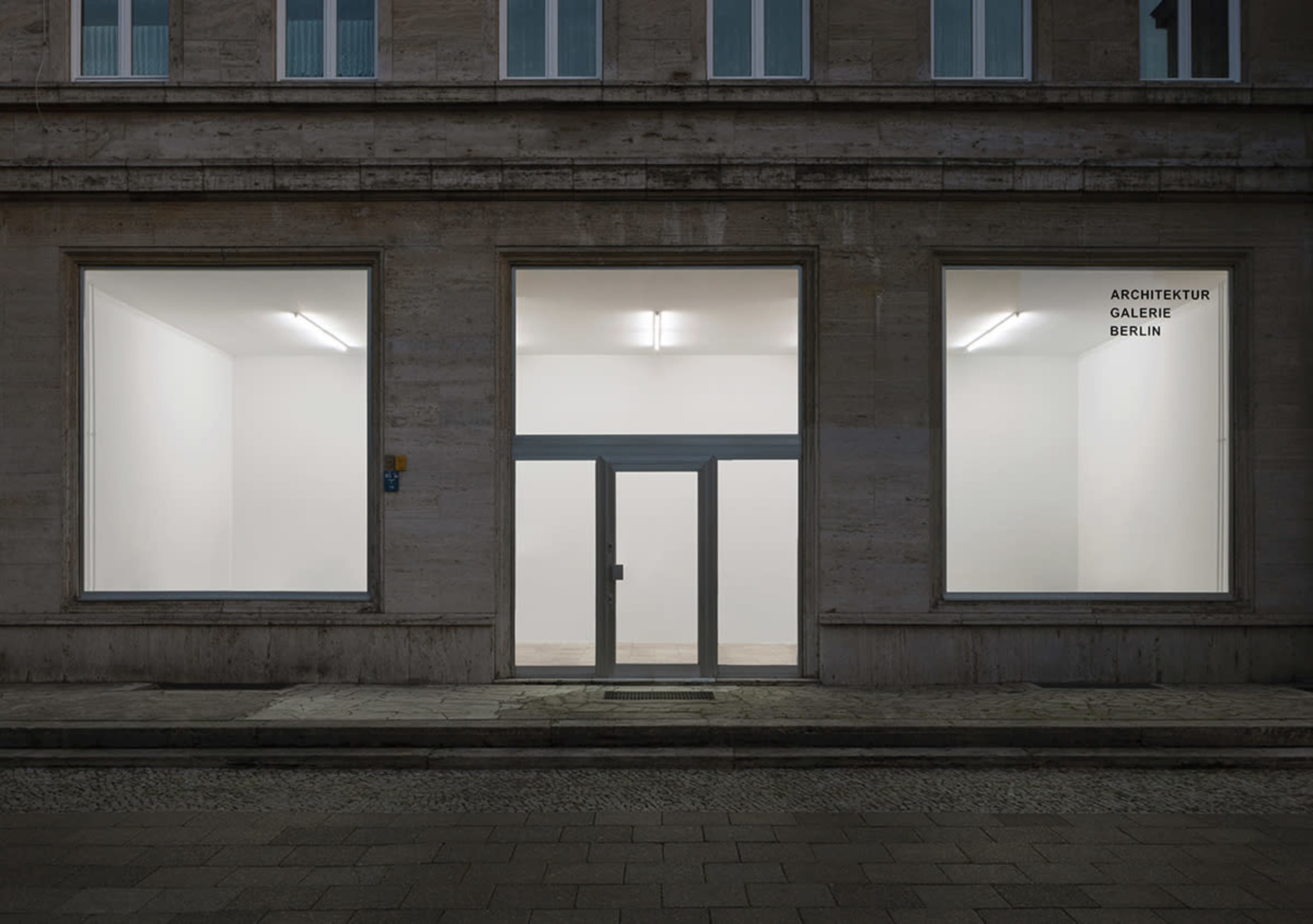 The exterior of a minimalist gallery space with large windows, a central entrance, and plain walls, labeled "ARCHITEKTUR GALERIE BERLIN."