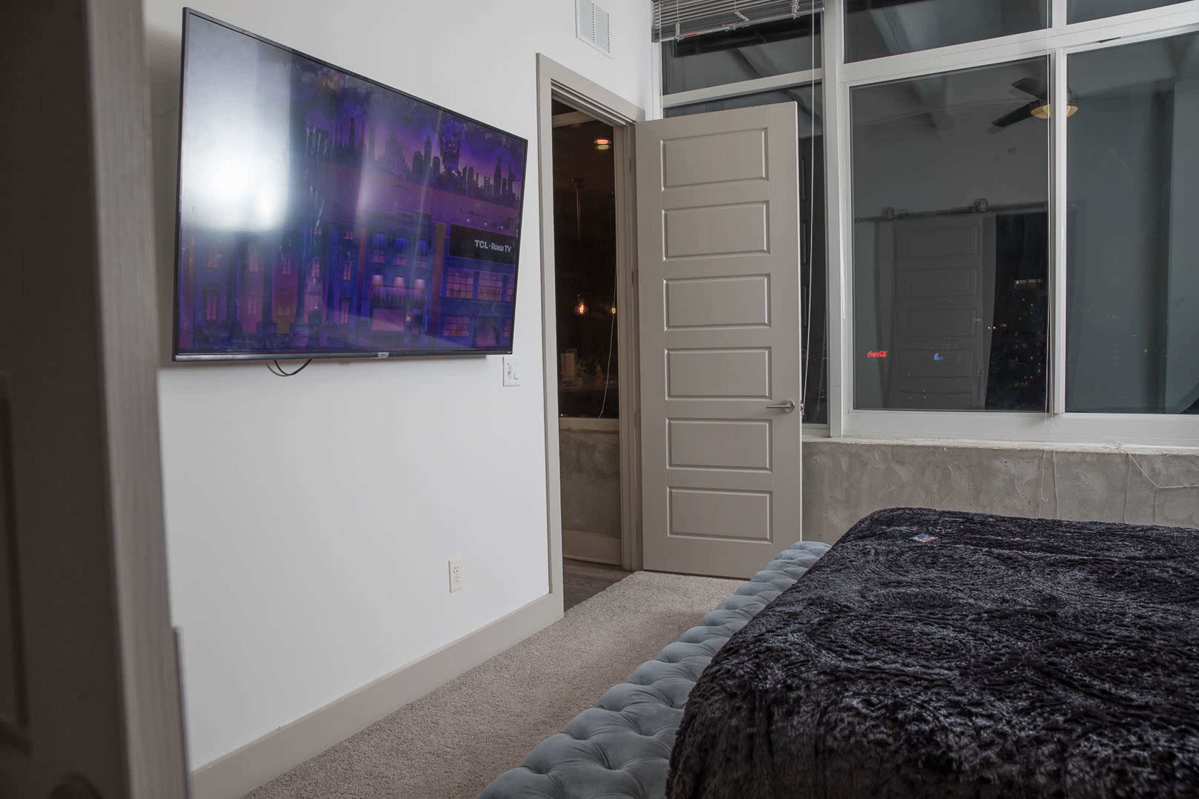 A bedroom with a large flat-screen TV mounted on the wall, a gray upholstered bed, and an open door leading to another area.
