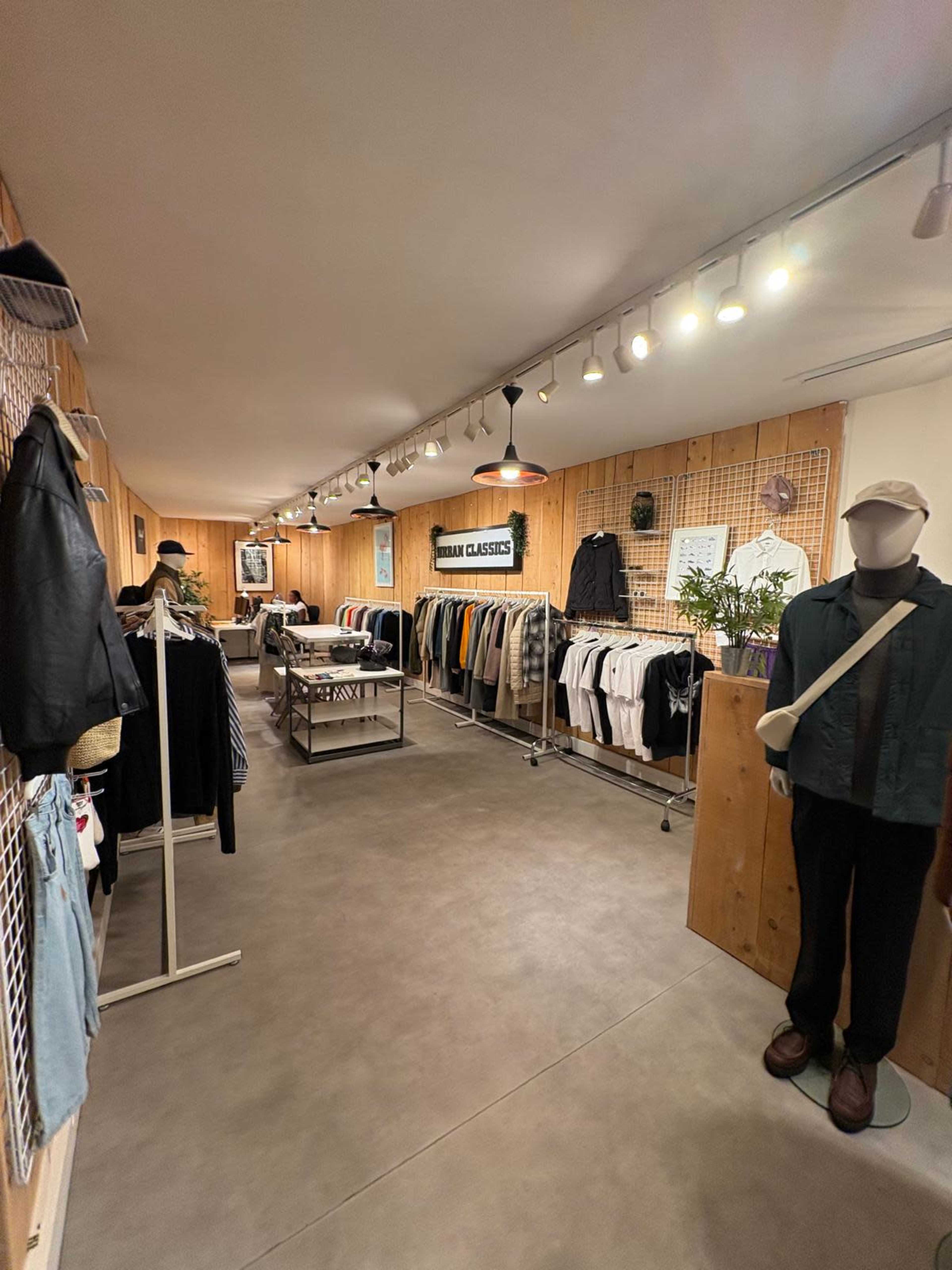 The image shows a clothing store interior with racks of apparel on either side, wooden walls, and a central display table under overhead lighting.