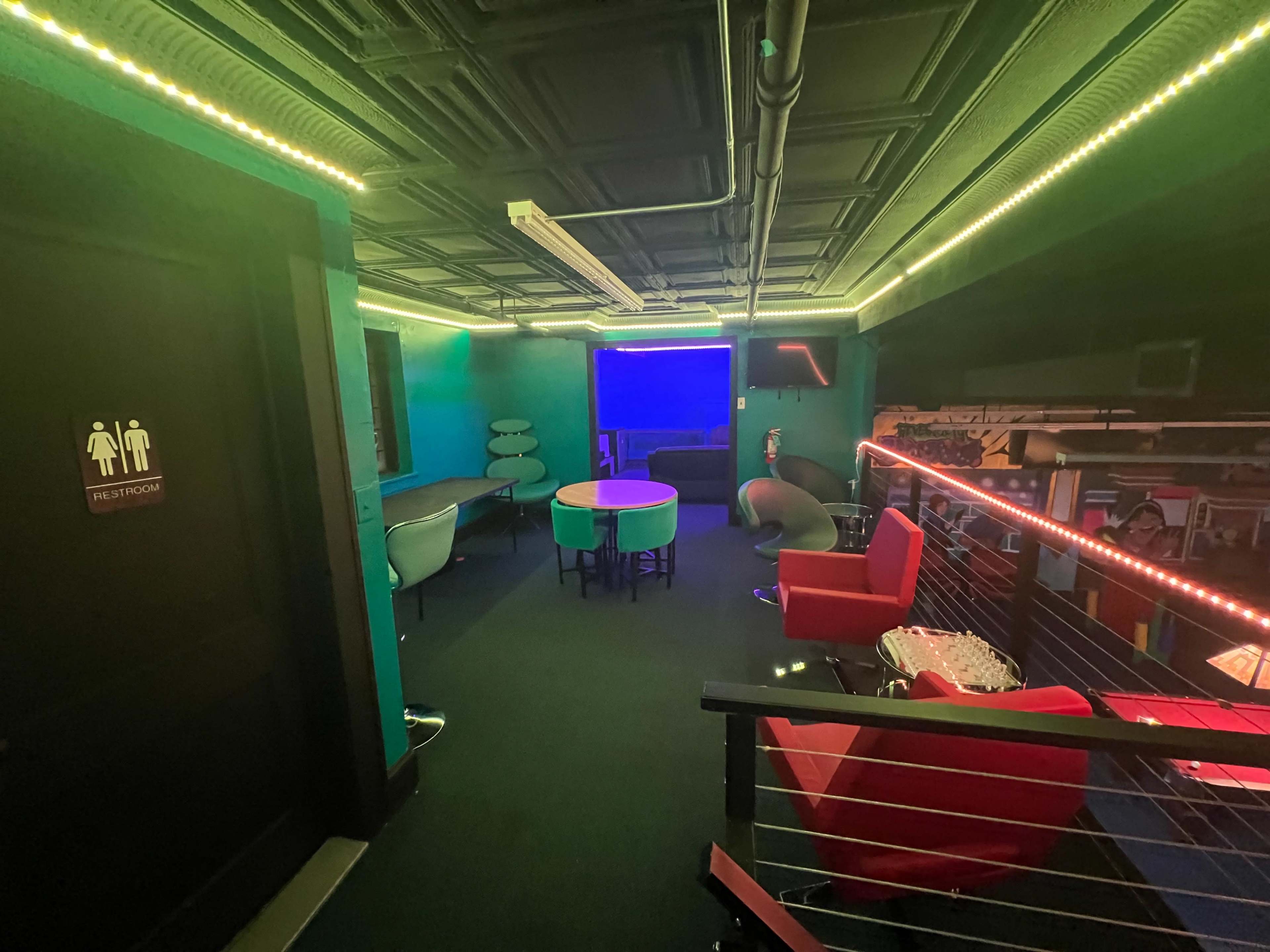 A brightly lit lounge area with colorful walls, a green and white table in the center, and various seating options including red chairs and a dark partition marking a restroom.