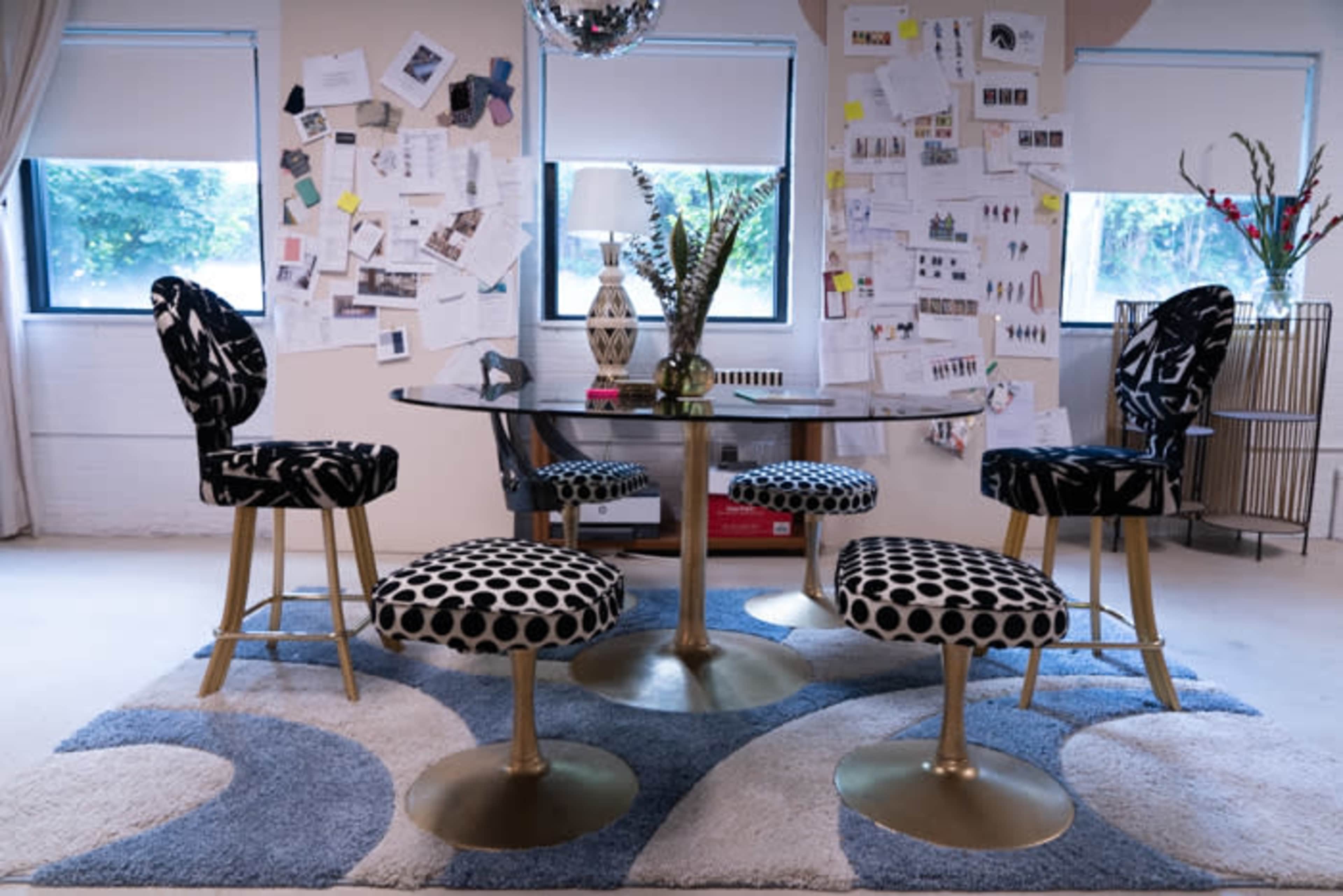 A dining area with a glass table surrounded by upholstered chairs and a patterned rug, against a backdrop of a wall covered with various notes and sketches.