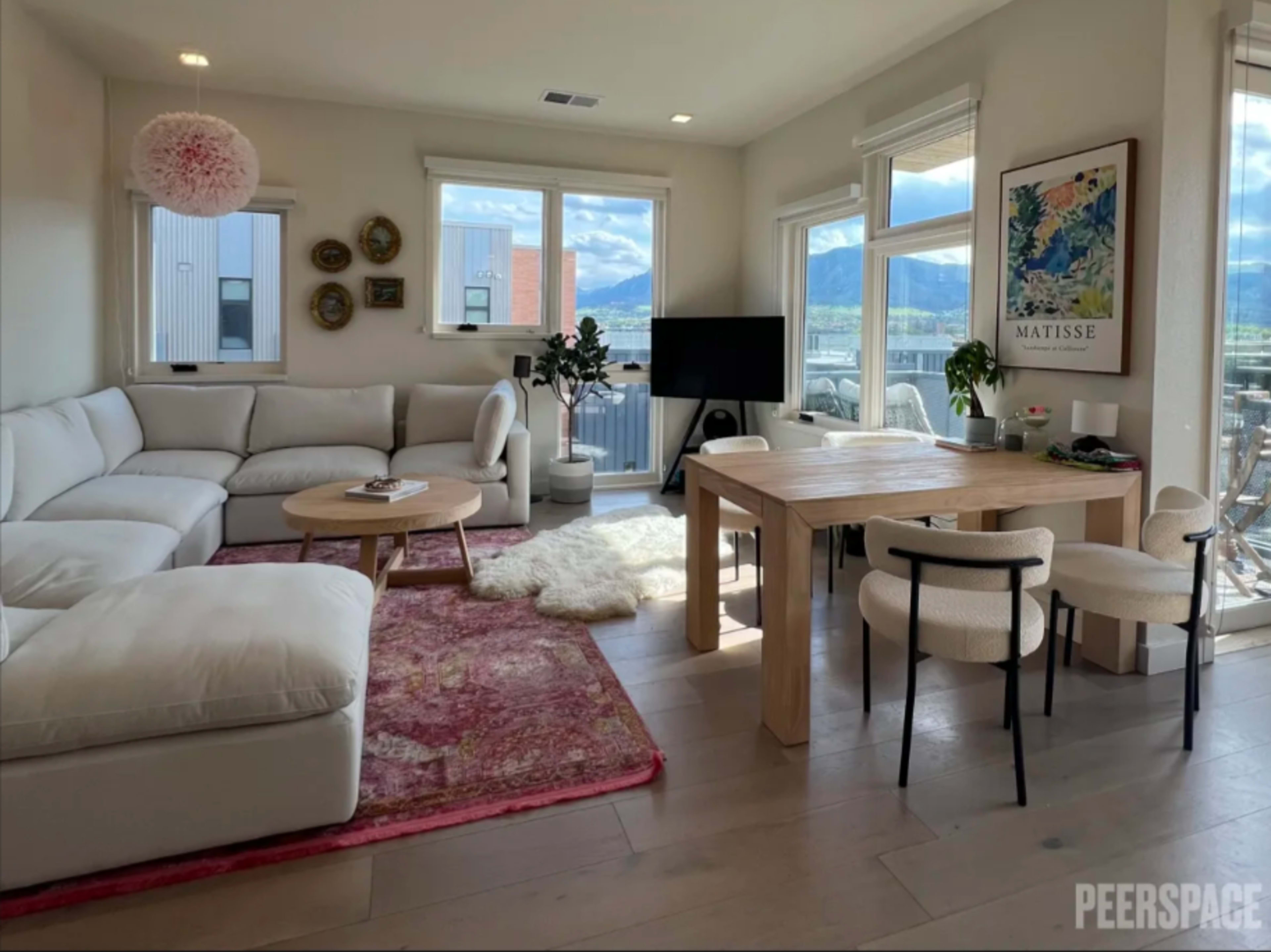 A modern living room with a sectional sofa, a wooden dining table, and large windows that provide a view of the outdoors.
