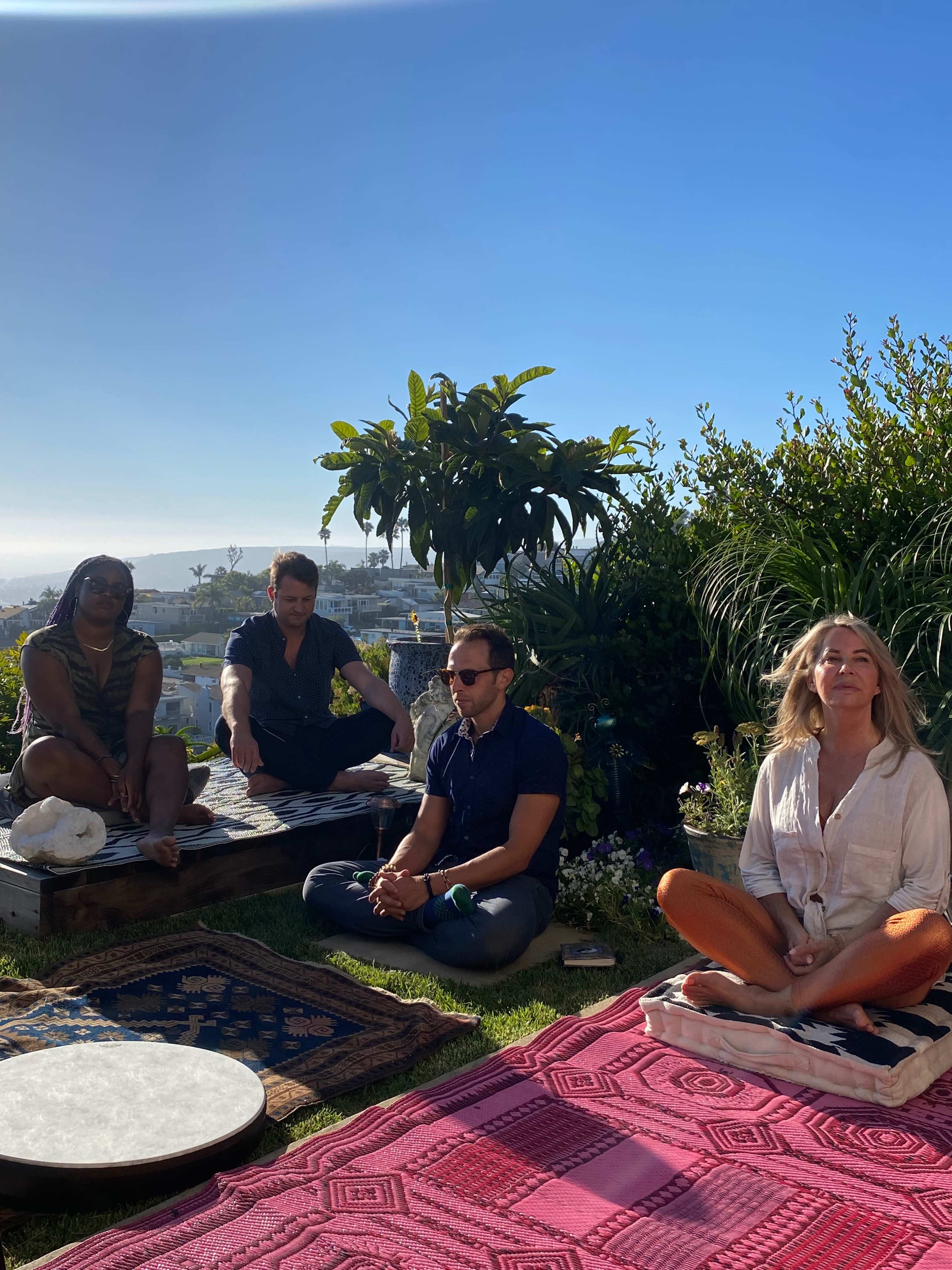 A group of four people sits on colorful mats in a sunny outdoor setting surrounded by plants.