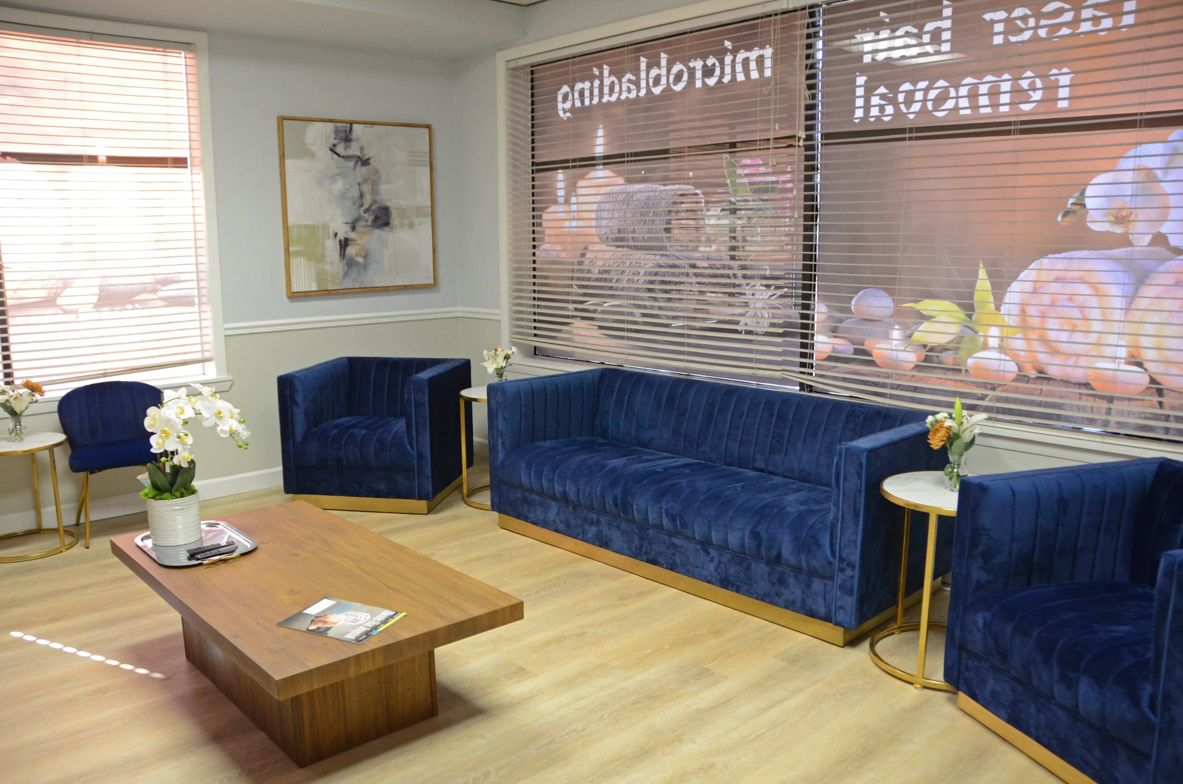 A waiting area with blue velvet chairs and a sofa, a wooden coffee table, and large windows featuring a decorative display.