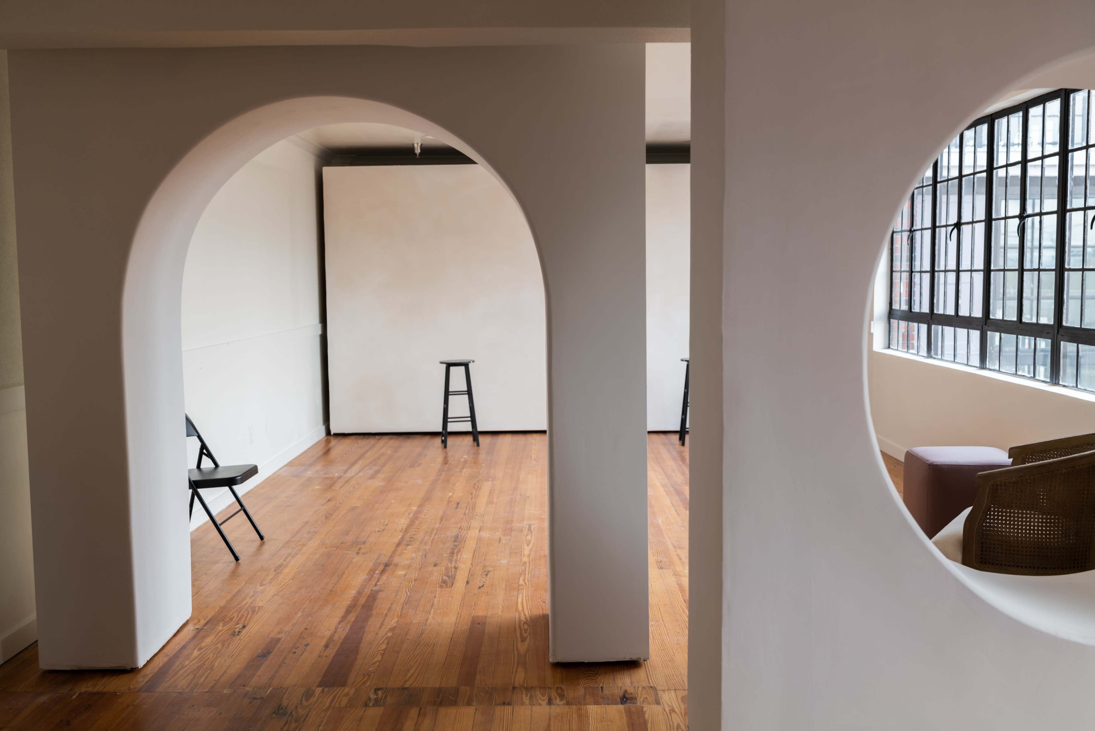 A minimalist interior space with arched openings and wooden floors, featuring two black stools placed in the room.
