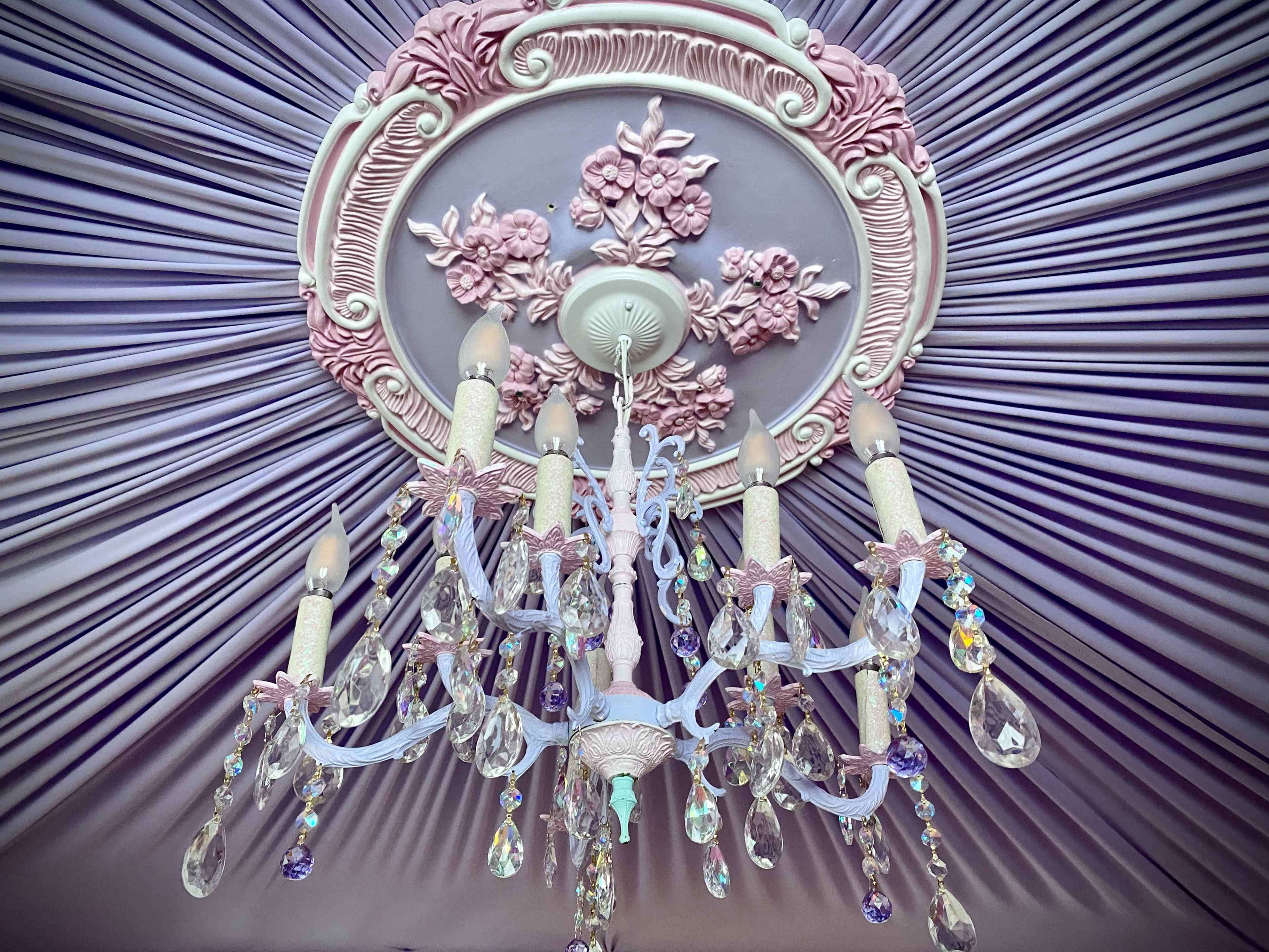 An ornate chandelier with multi-colored crystals suspended from a decorative ceiling medallion featuring floral designs, all set against a pleated pastel-colored ceiling.