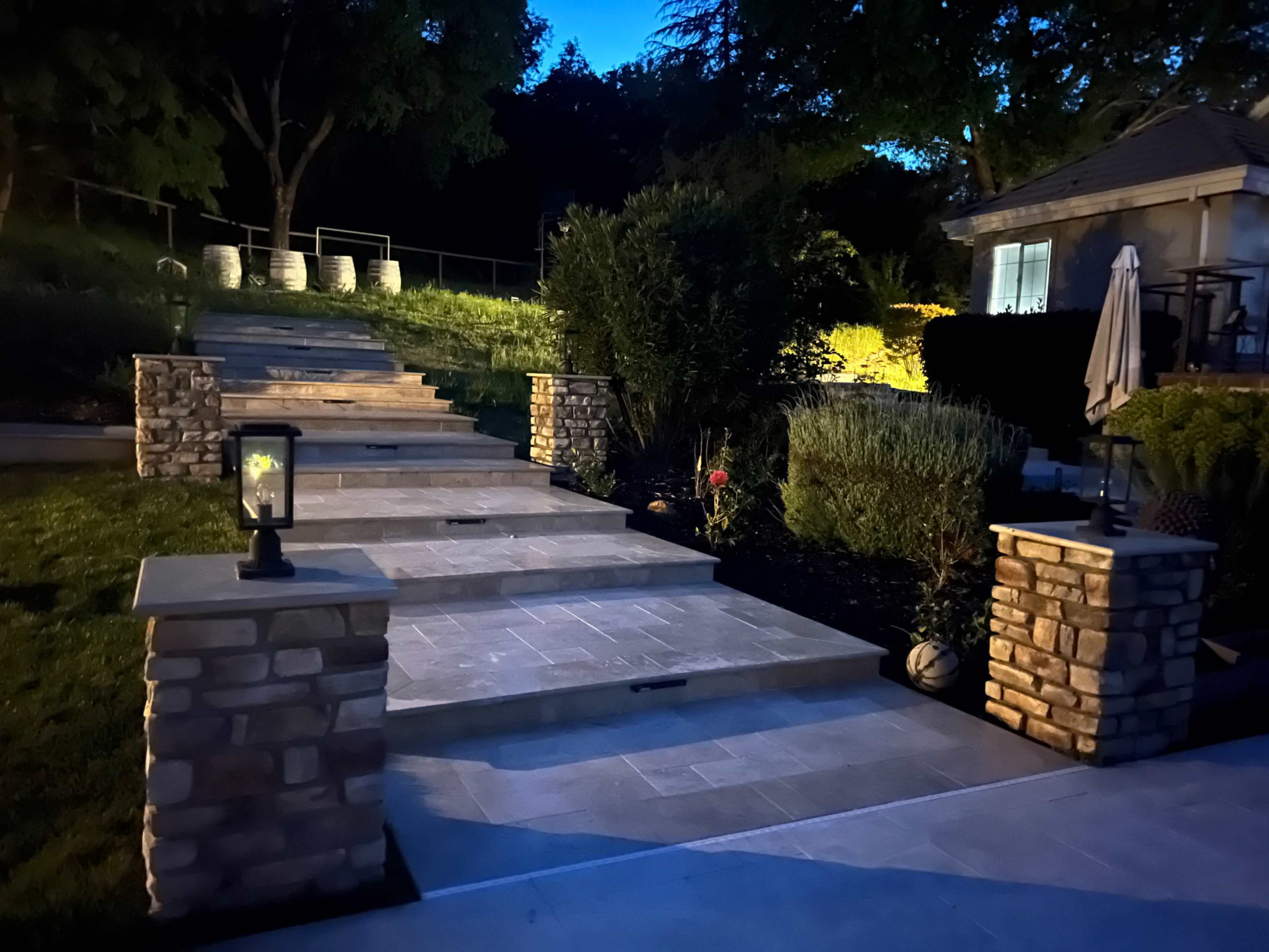 The image shows a stone pathway illuminated by pathway lights, bordered by gardens and leading to a house at dusk.