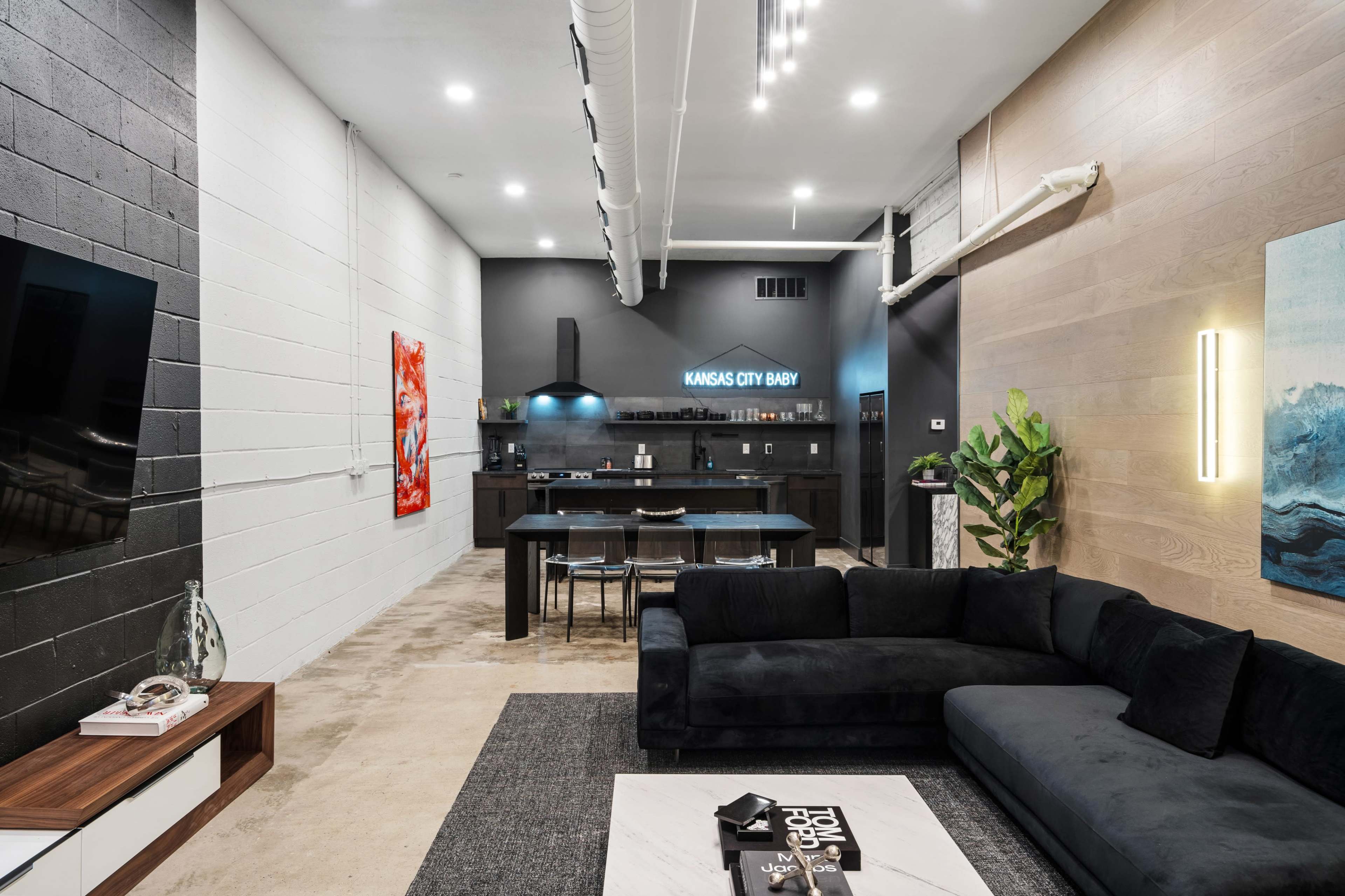 The image shows a modern, stylish living space featuring a black sectional sofa, a coffee table, and an open kitchen at the far end with sleek cabinetry.