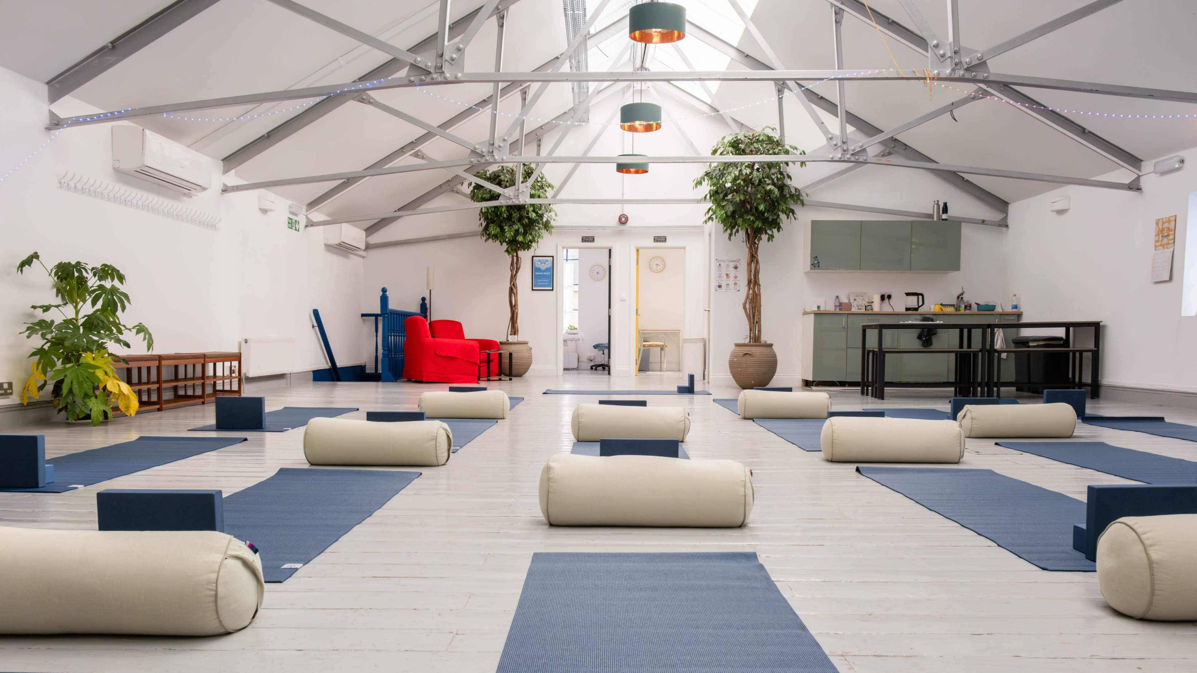 The image shows a spacious yoga studio with rolled mats and bolsters arranged neatly on a wooden floor beneath a high ceiling with hanging lights.