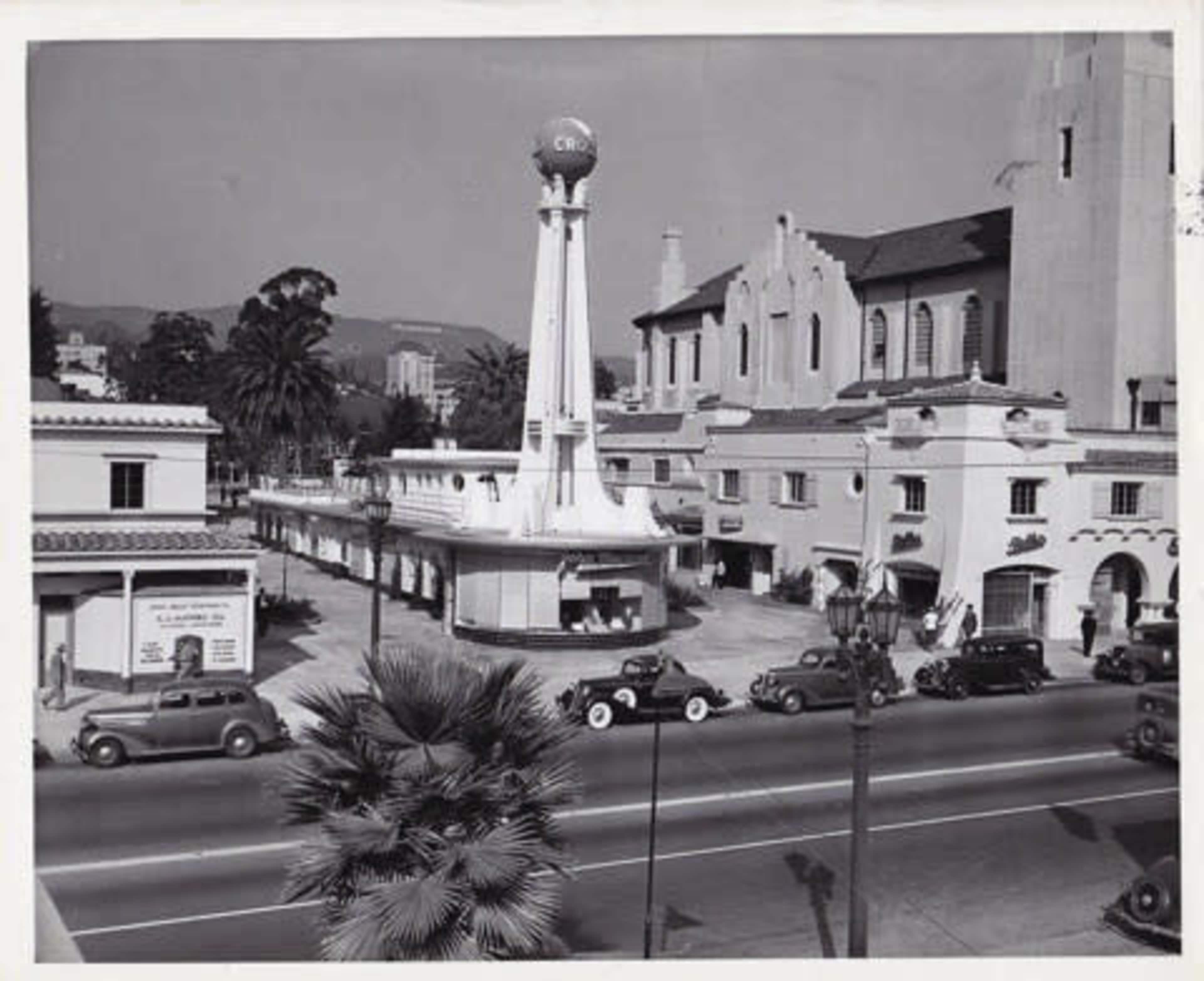 A historic black-and-white photograph depicts a street scene with vintage cars parked in front of a building featuring a tall tower and signage, surrounded by palm trees.