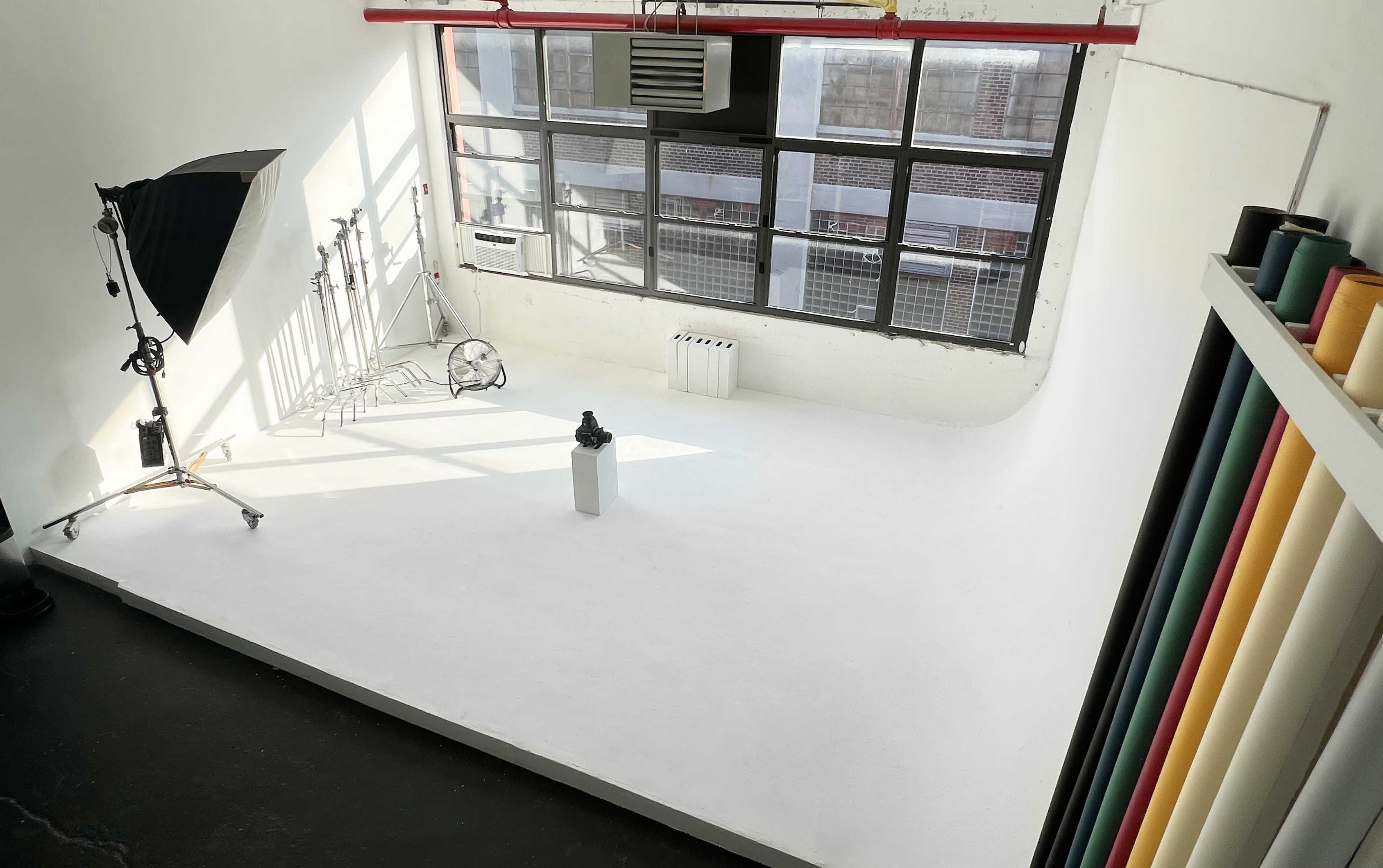 The image shows a well-lit photography studio with a white backdrop, a lighting setup, and a variety of colorful rolls of backdrops arranged on the side.